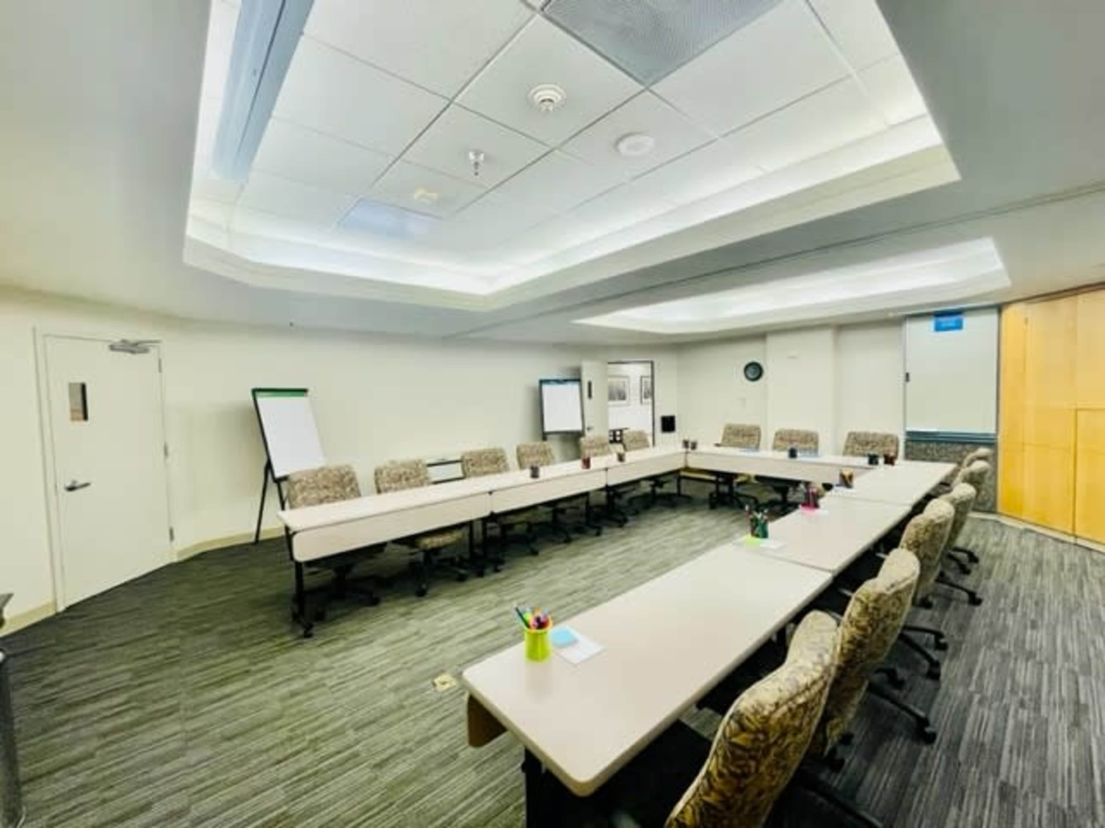 A conference room features a rectangular arrangement of tables and chairs, with a whiteboard and presentation screen at one end.