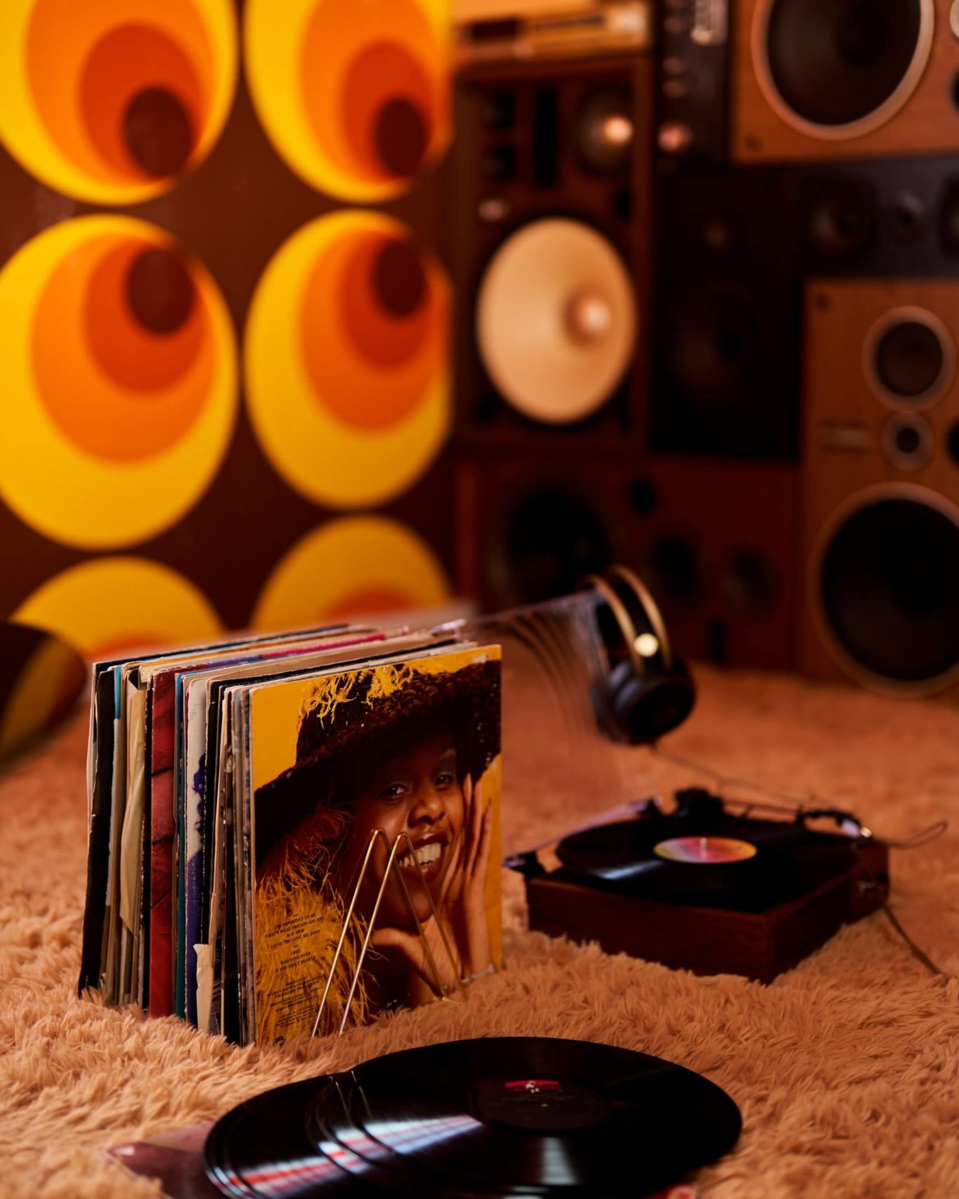 A collection of vinyl records and a turntable are set against a retro-themed wall and large speakers in a cozy room.