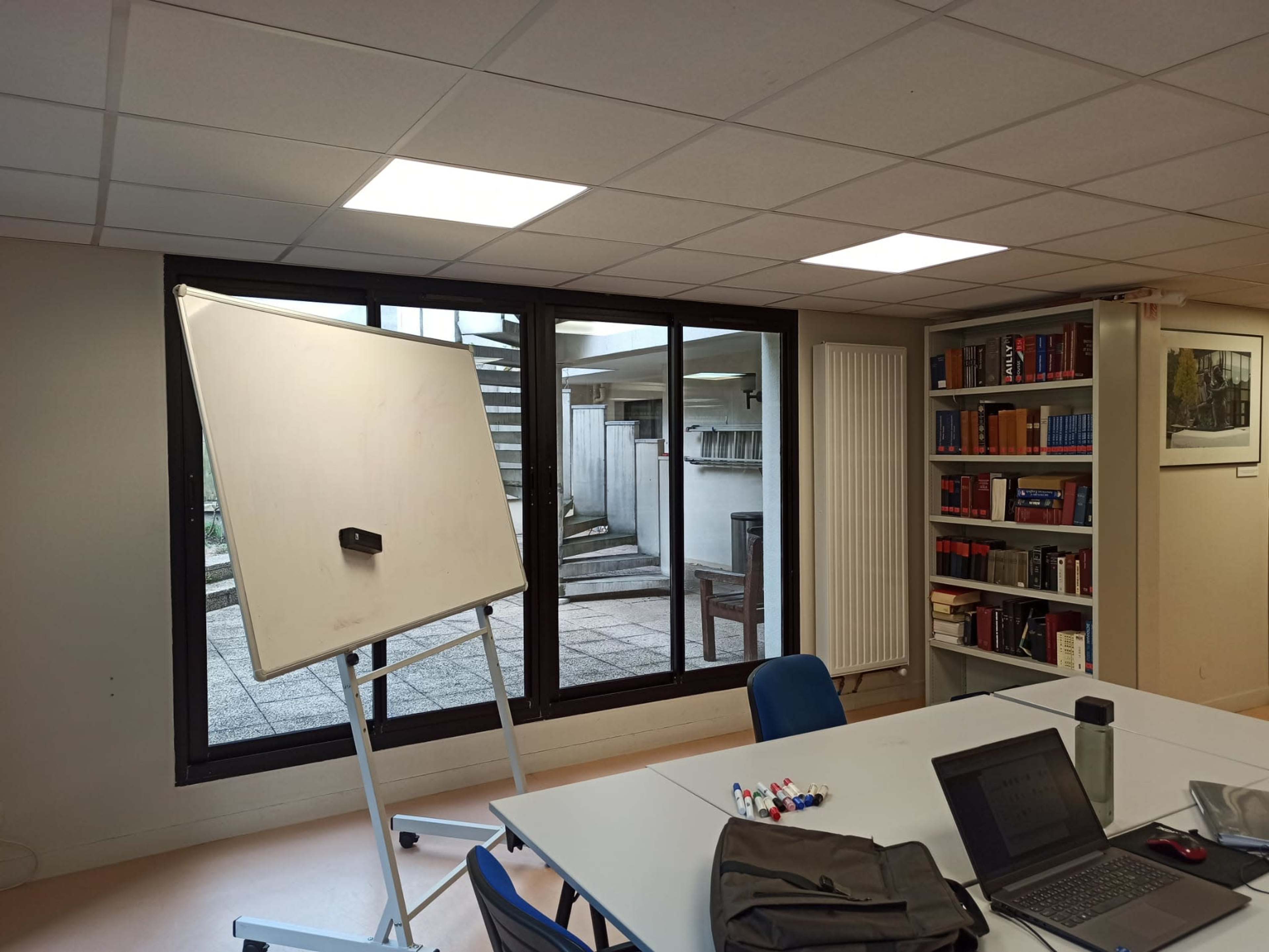 A meeting room features a whiteboard, a table with a laptop and markers, and large windows overlooking a staircase and outdoor area.