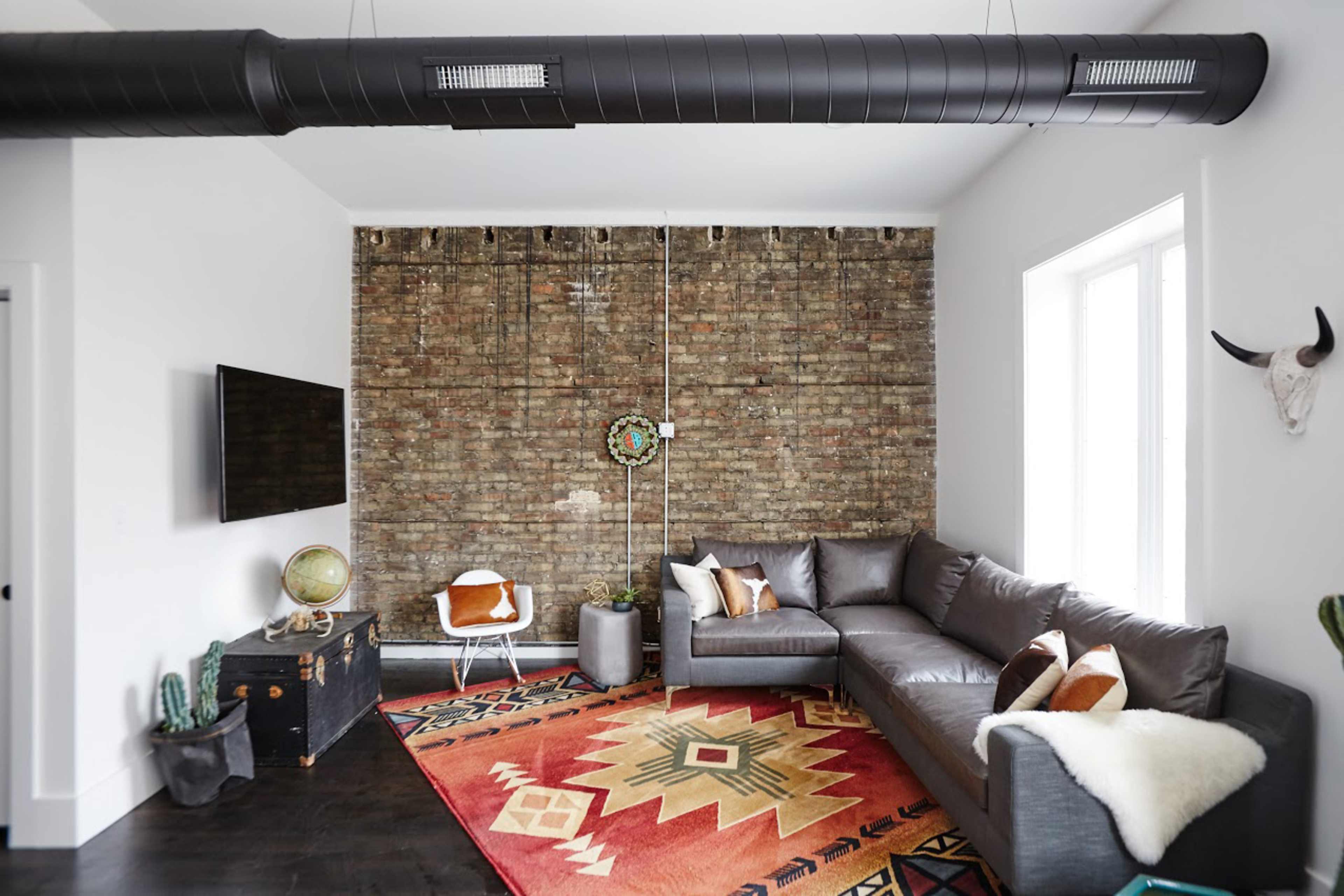 The living room features a sectional sofa, a patterned rug, a brick wall, and a television mounted on the opposite wall.