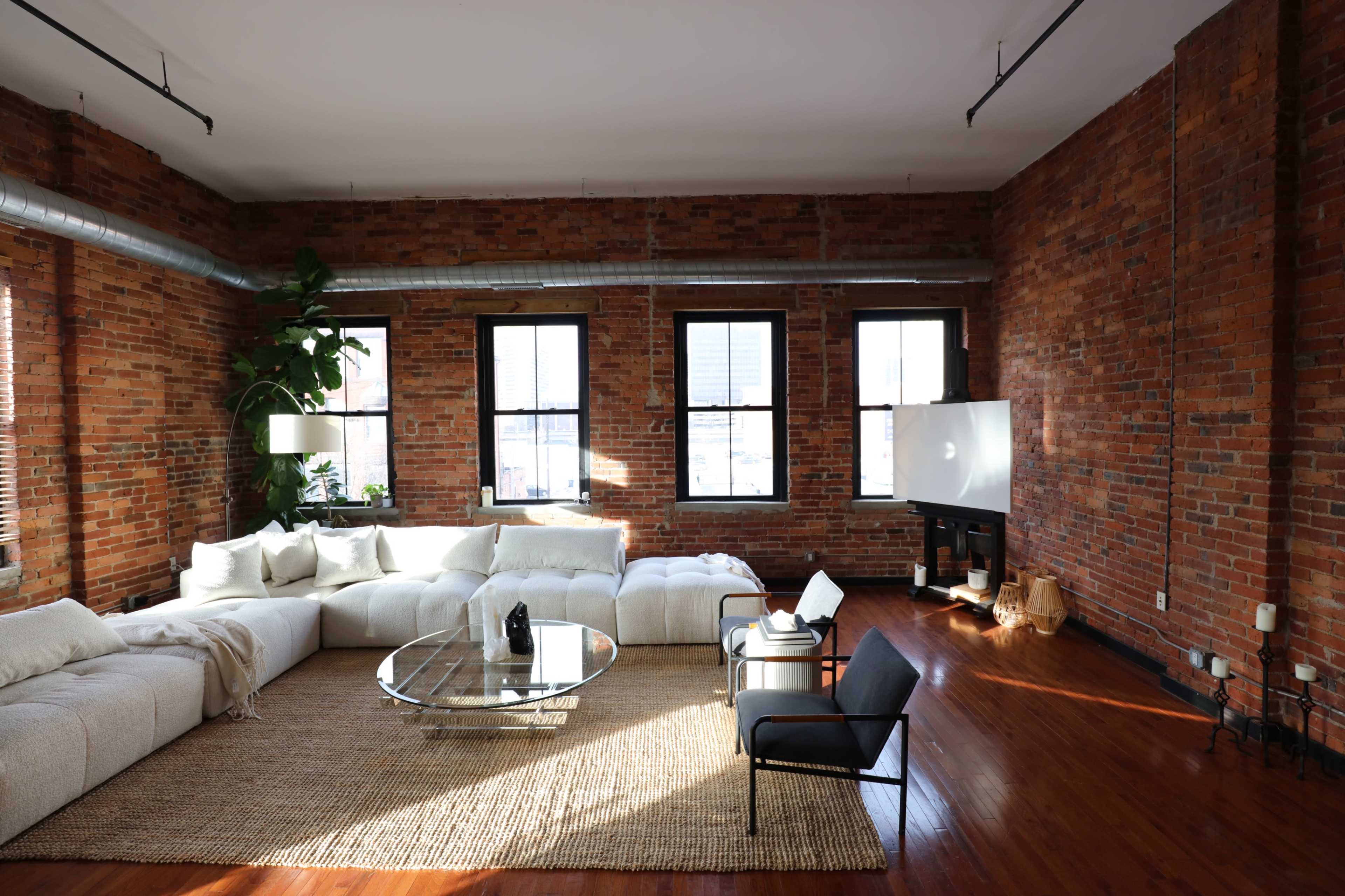 A spacious living room with a large sectional sofa, a glass coffee table, and a flat-screen TV against a brick wall.