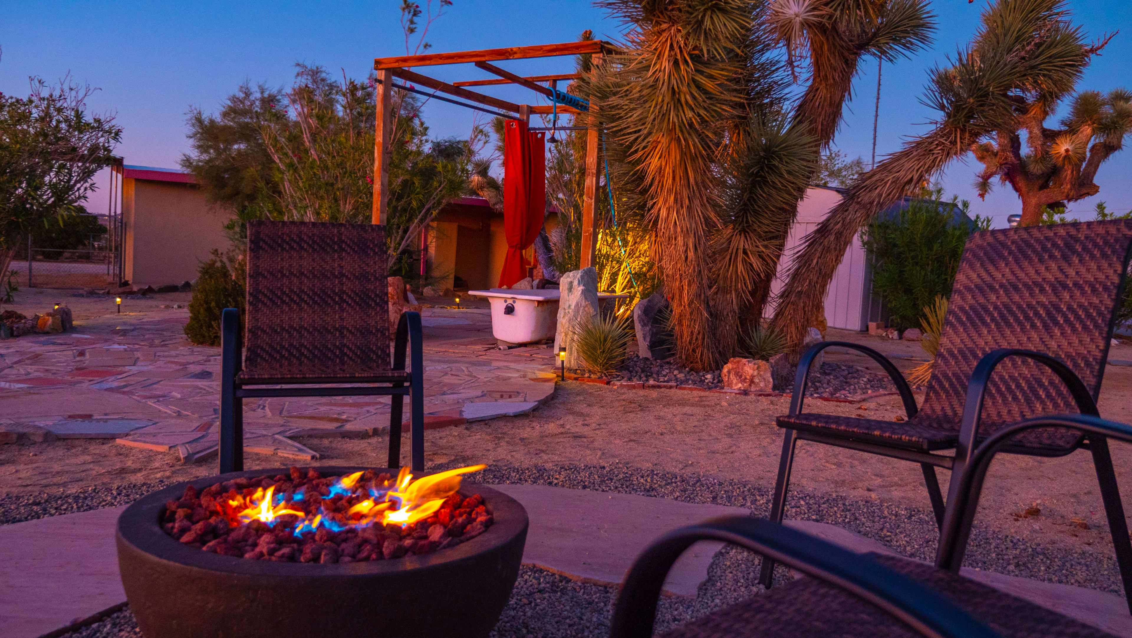 A fire pit with glowing flames is surrounded by two chairs in a desert garden with palm trees and a small structure in the background.