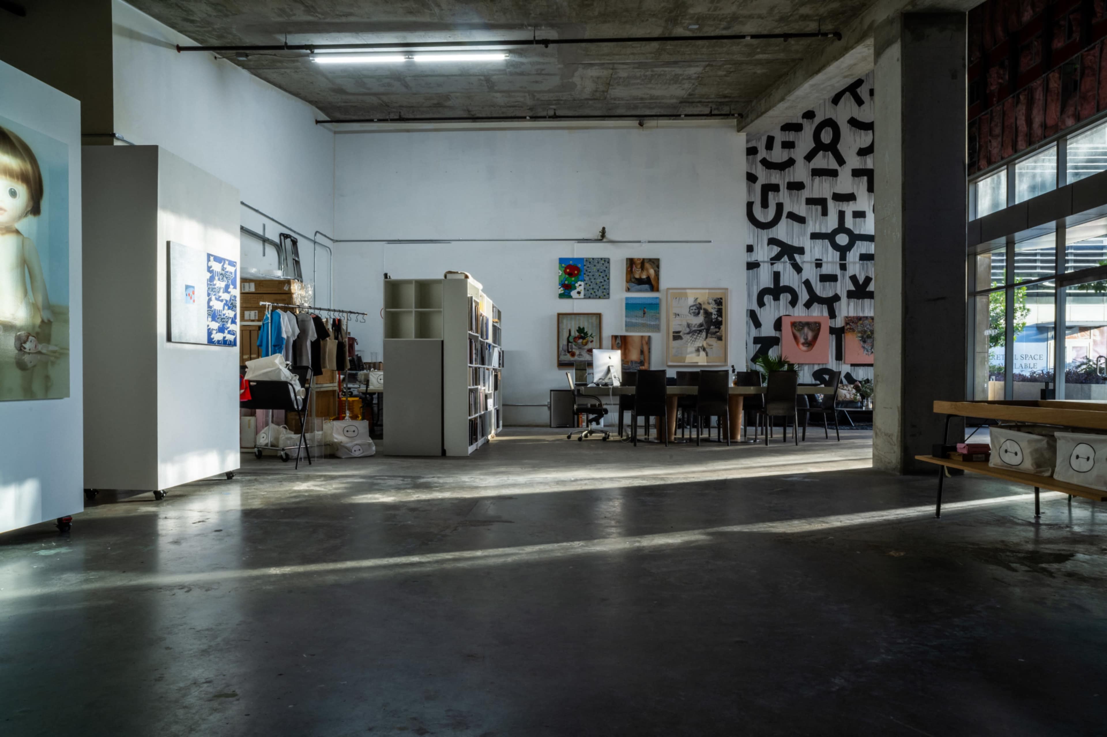 The image shows a spacious interior of an art gallery or studio with various artworks displayed on the walls and a few tables and chairs arranged for visitors.
