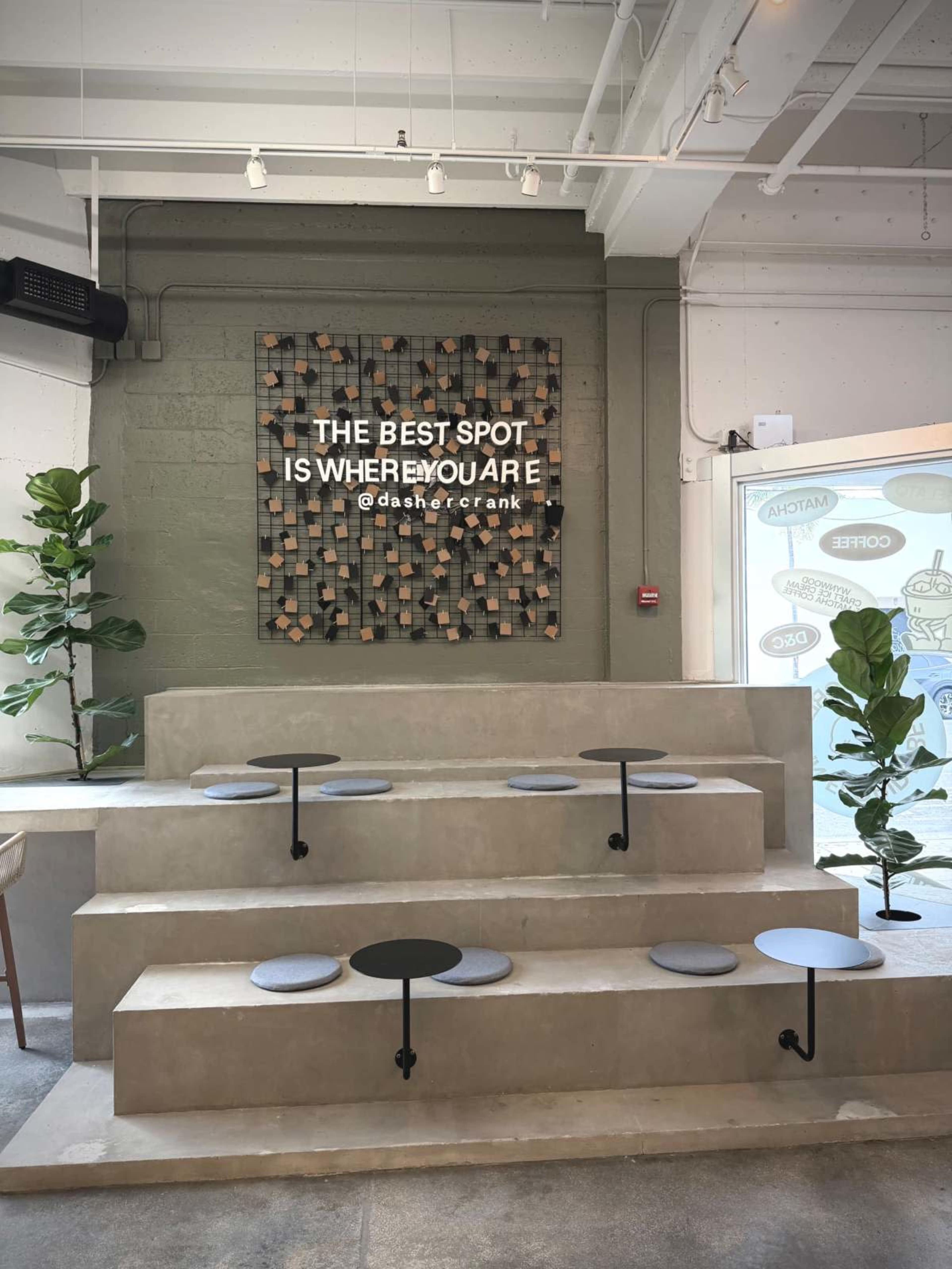 The image shows a modern seating area with tiered concrete steps and small round tables, complemented by green plants and a large decorative wall sign.