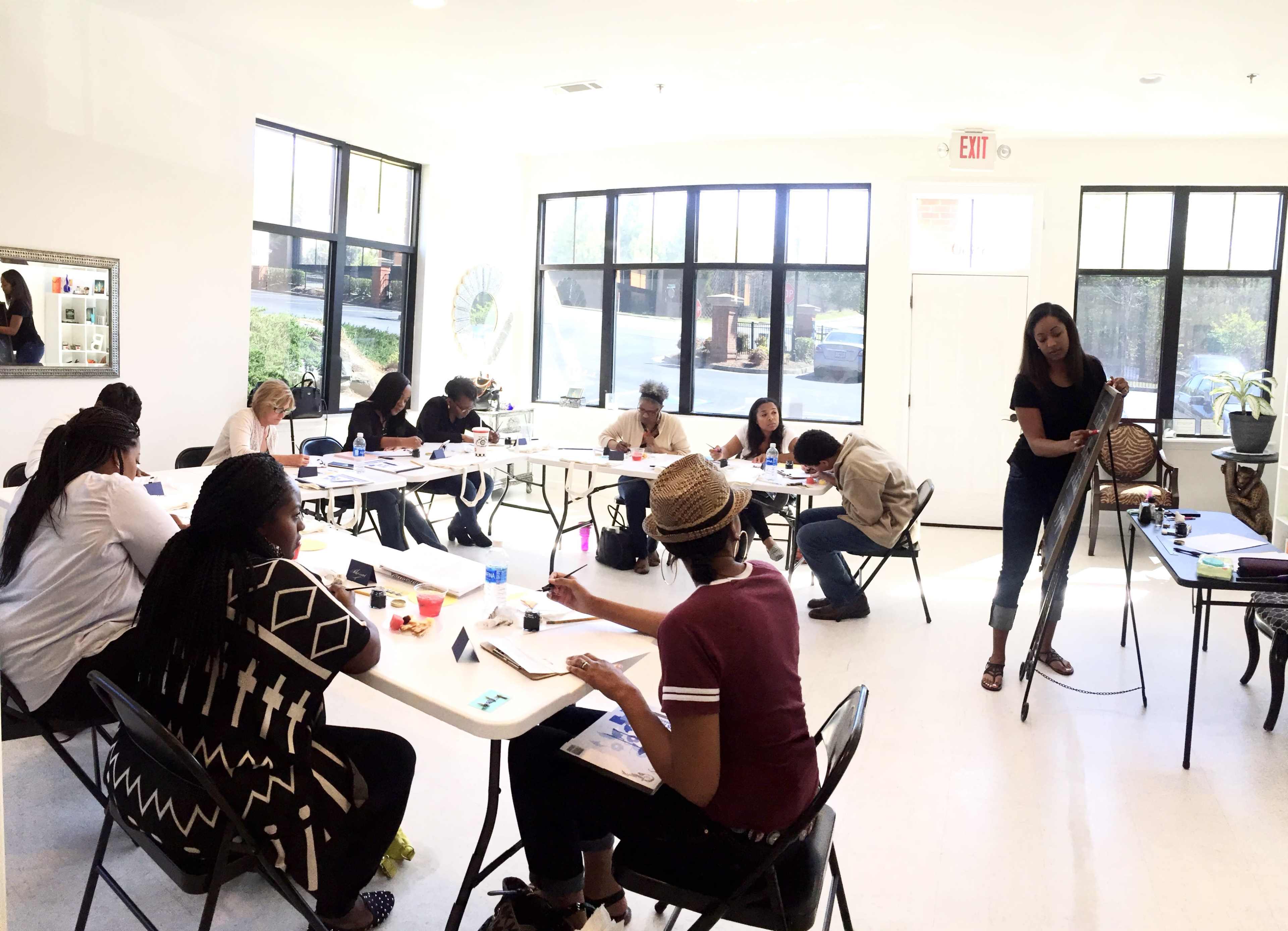 A group of people participates in an art class, sitting at tables with art supplies while one instructor demonstrates techniques.