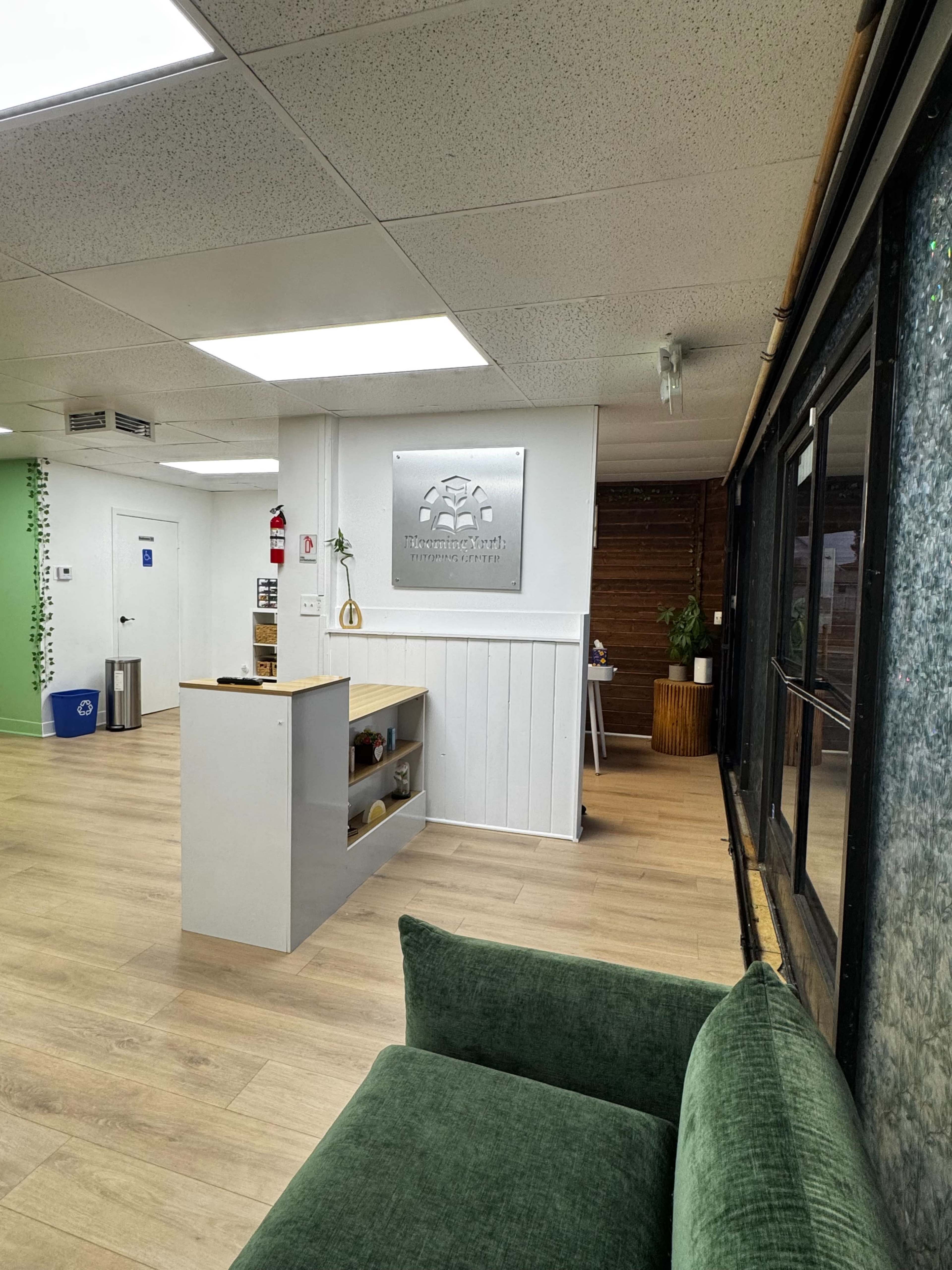 The image shows a modern office interior with a reception area featuring a wooden counter, a logo sign on the wall, and green decor elements, including a plant.