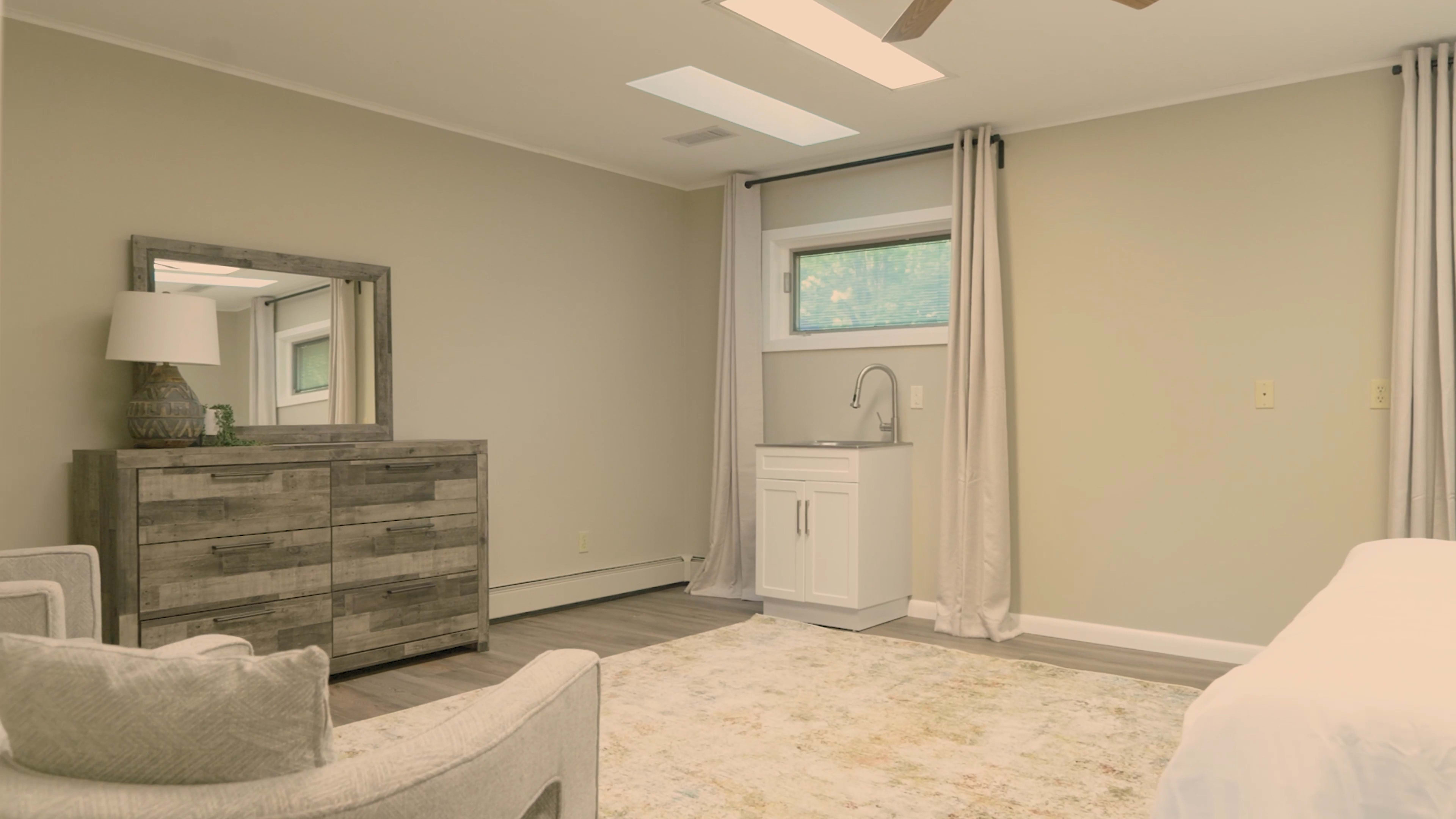 The image shows a light-colored room with a dresser, a mirror, a small kitchenette area, and a window with curtains.
