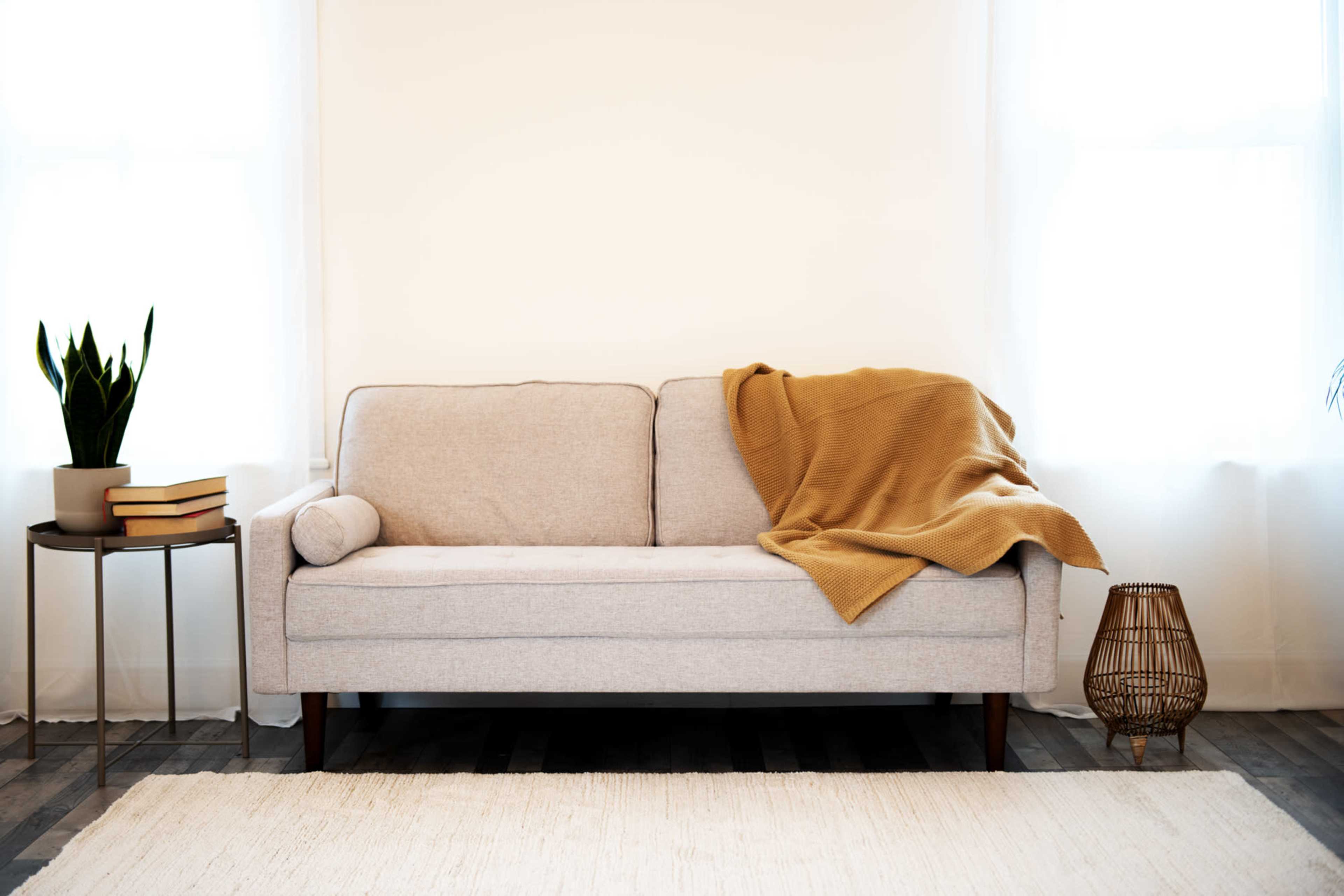 A light gray sofa with a mustard-colored blanket draped over it sits against a white wall, accompanied by a small side table with books and a potted plant.