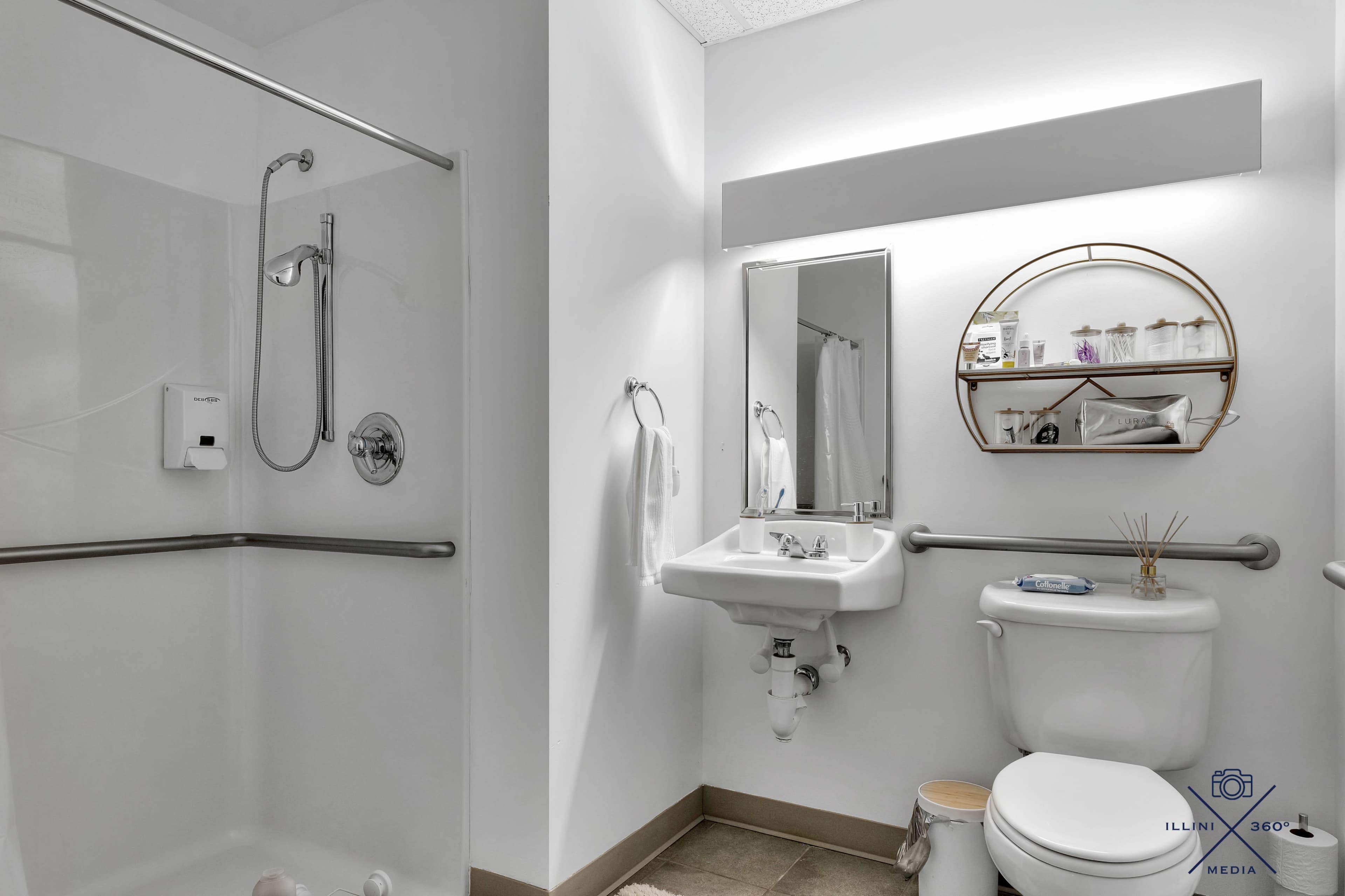 The image shows a bathroom featuring a shower stall, a sink with a mirror above, and a toilet with grab bars for assistance.