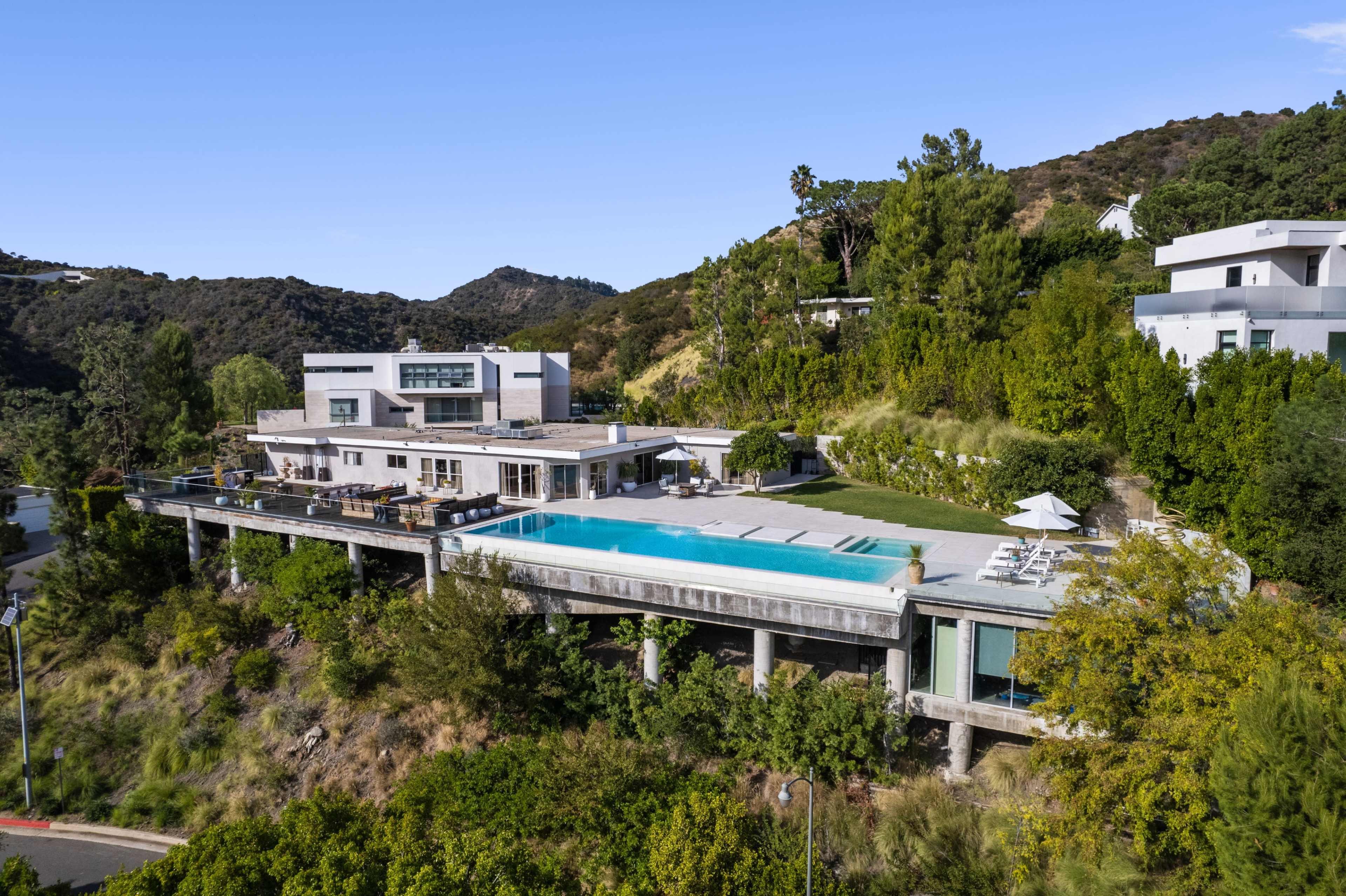 A modern house with a large swimming pool set on a hillside surrounded by greenery and mountains.