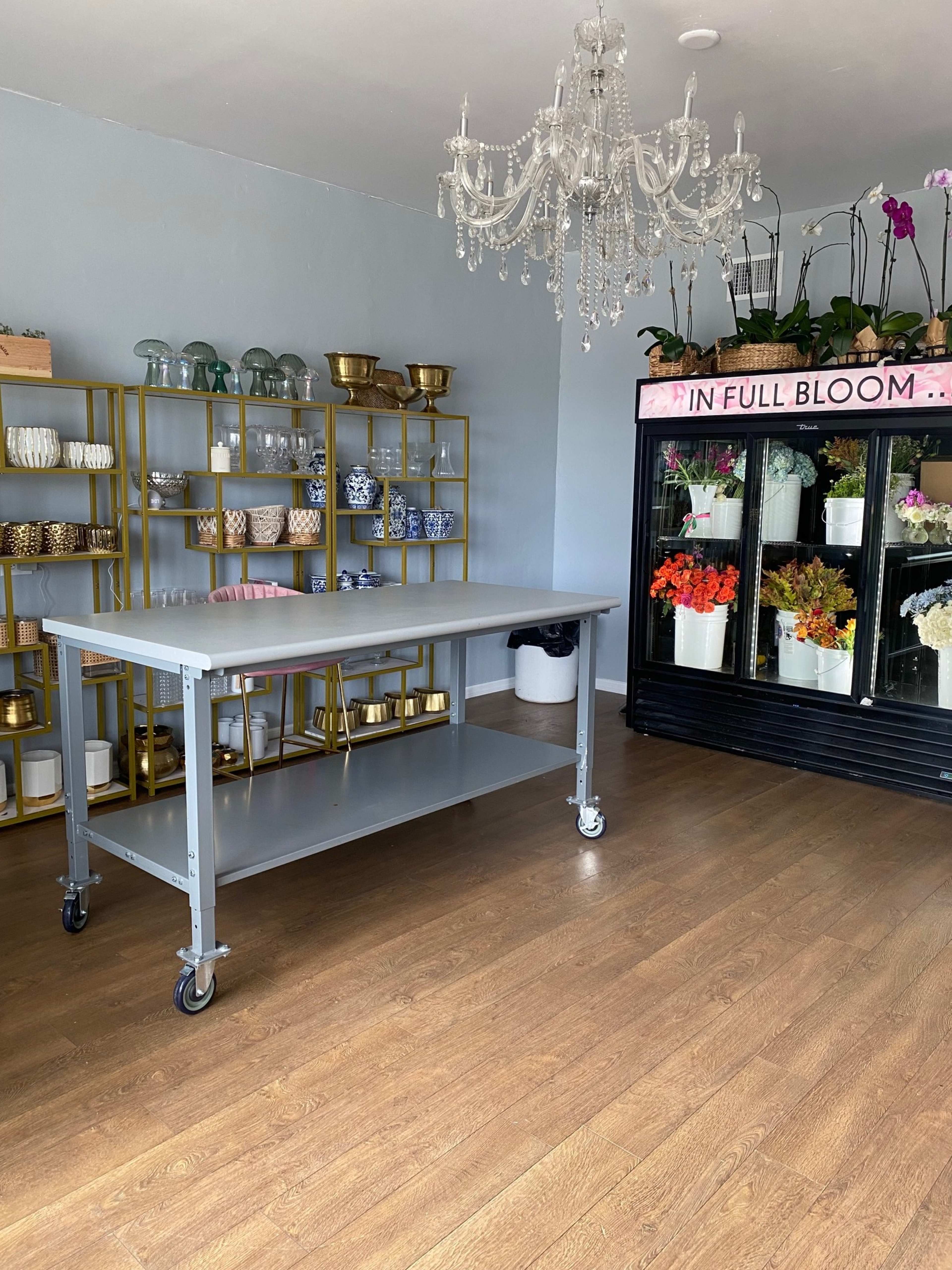 The image shows a well-lit room with a gray worktable on wheels, a gold-framed shelving unit filled with various glassware and decorative bowls, and a floral display cooler.