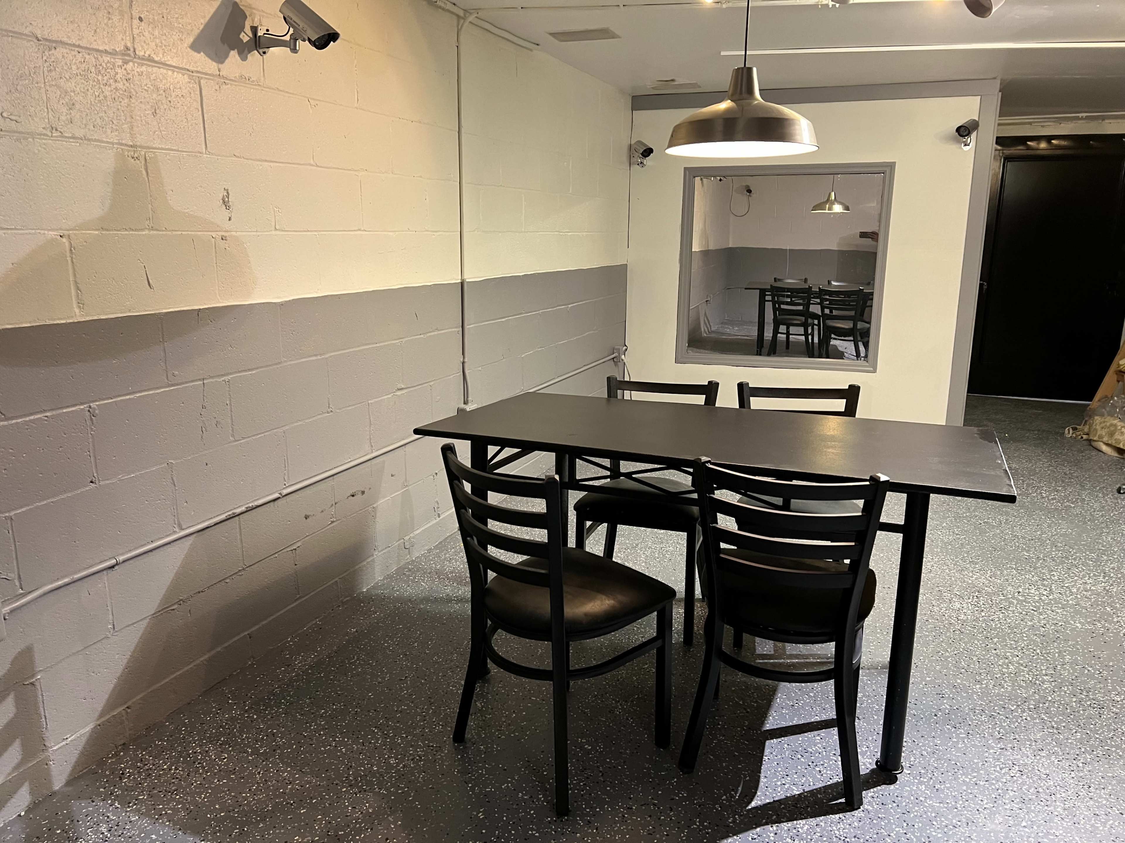 A black table with four chairs is positioned in a sparsely furnished room with gray walls and a pendant light overhead.