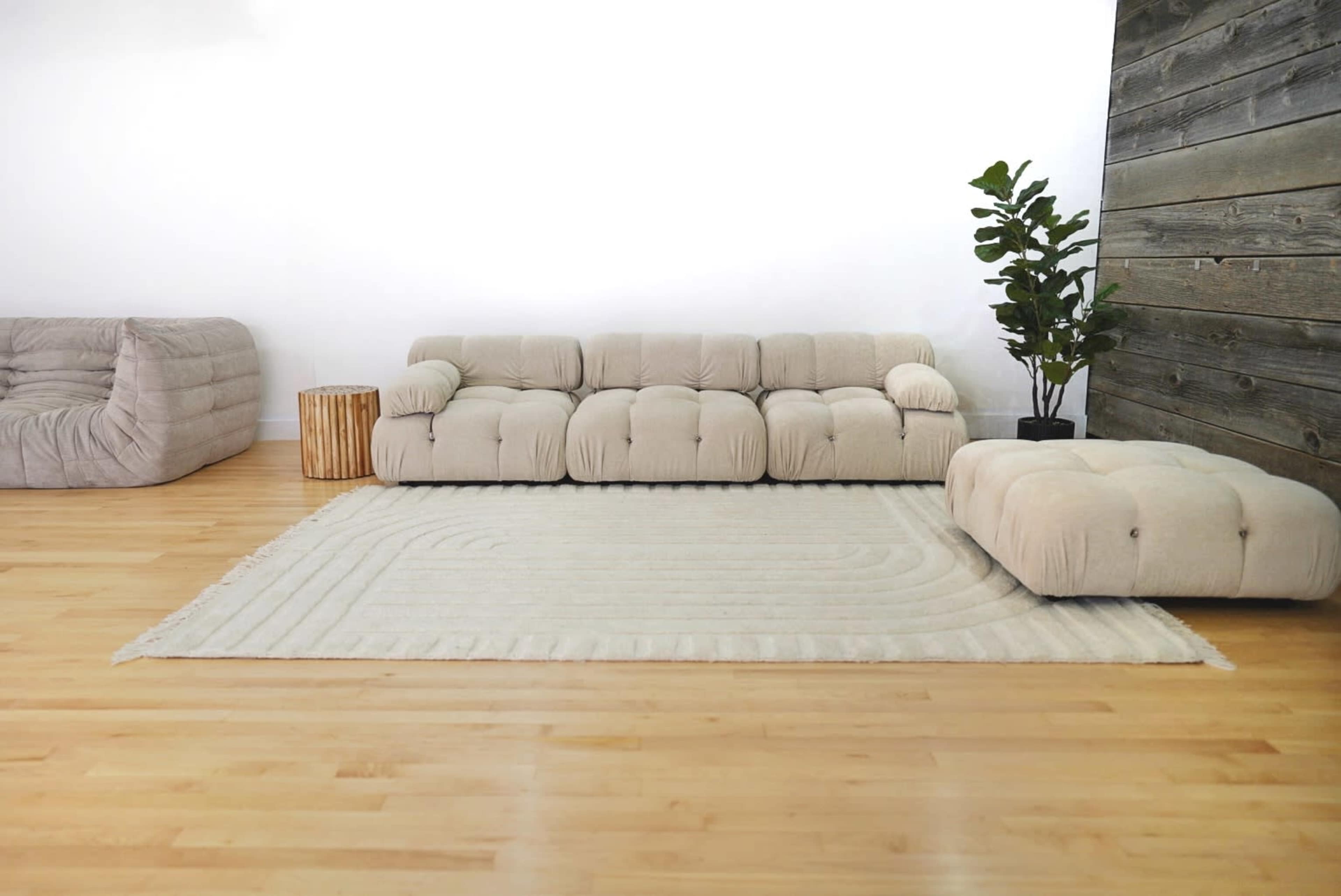 A minimalist living room featuring a beige, tufted sofa, a matching ottoman, a textured area rug, and a potted plant against a wooden accent wall.