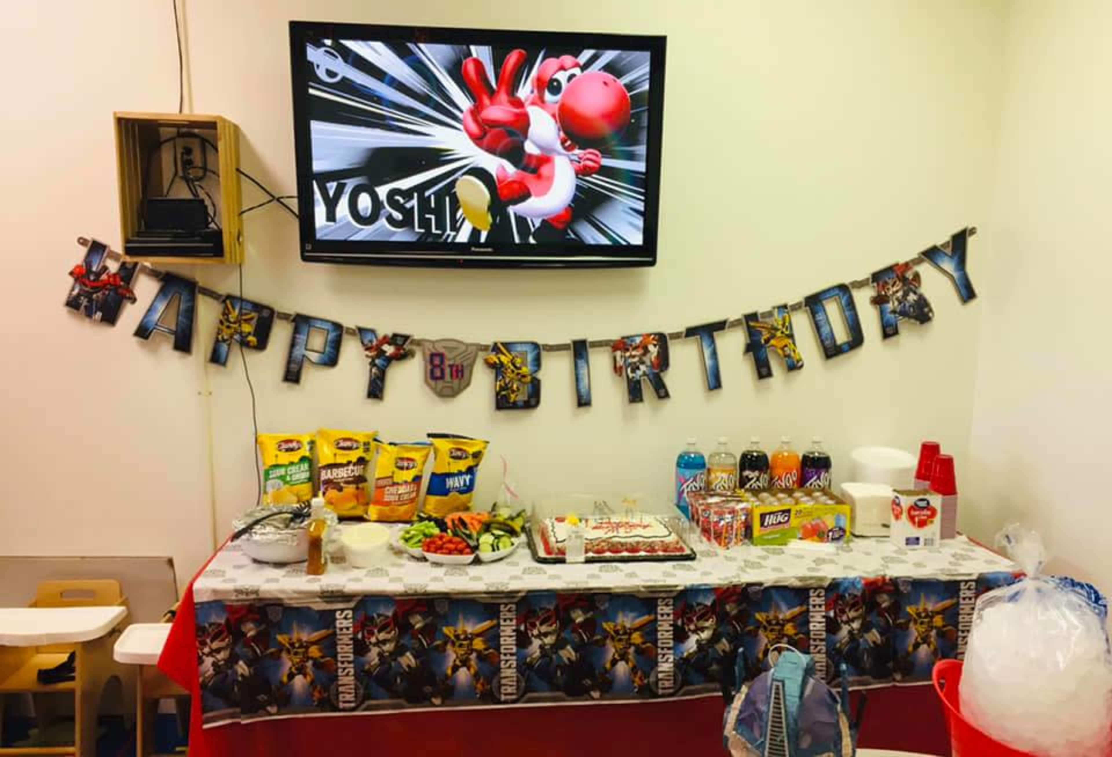 A birthday celebration setup with a table displaying snacks, drinks, and a "Happy 8th Birthday" banner beneath a television screen featuring a gaming character.
