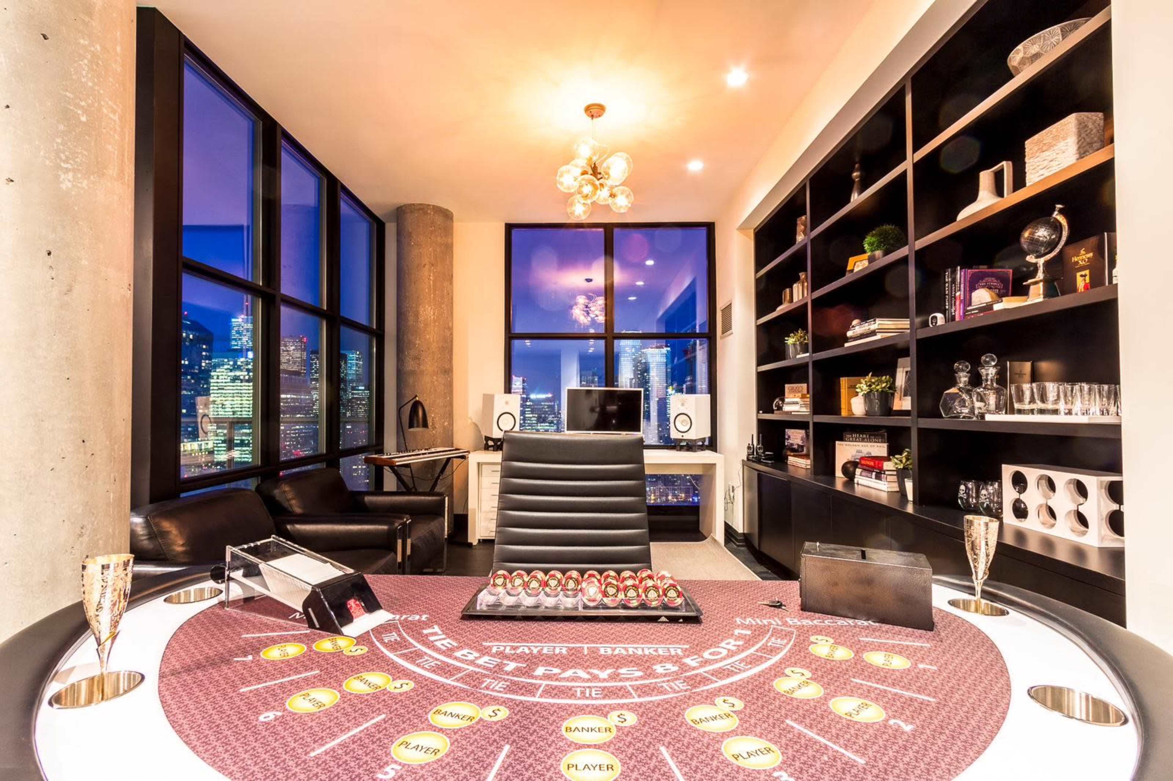 The image shows a modern game room with a casino-style table at the center, surrounded by large windows showcasing a city skyline at dusk.