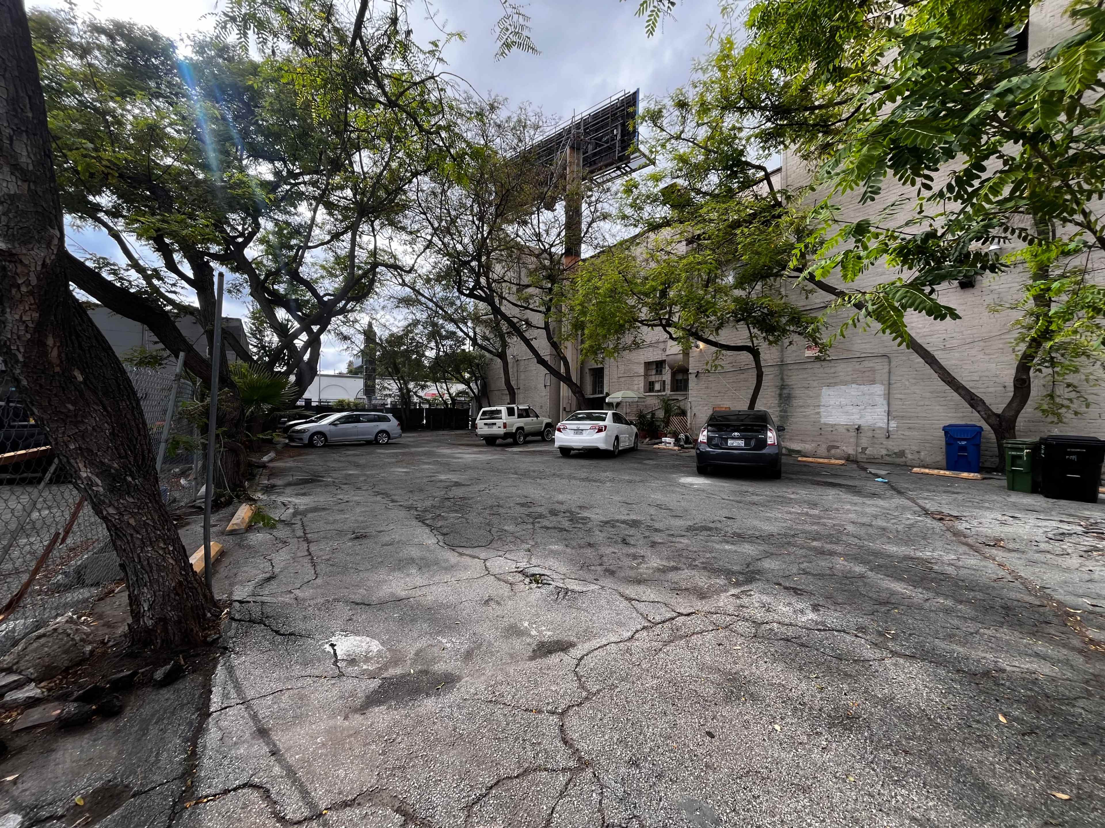 The image shows a vacant paved parking lot surrounded by trees and a brick building.