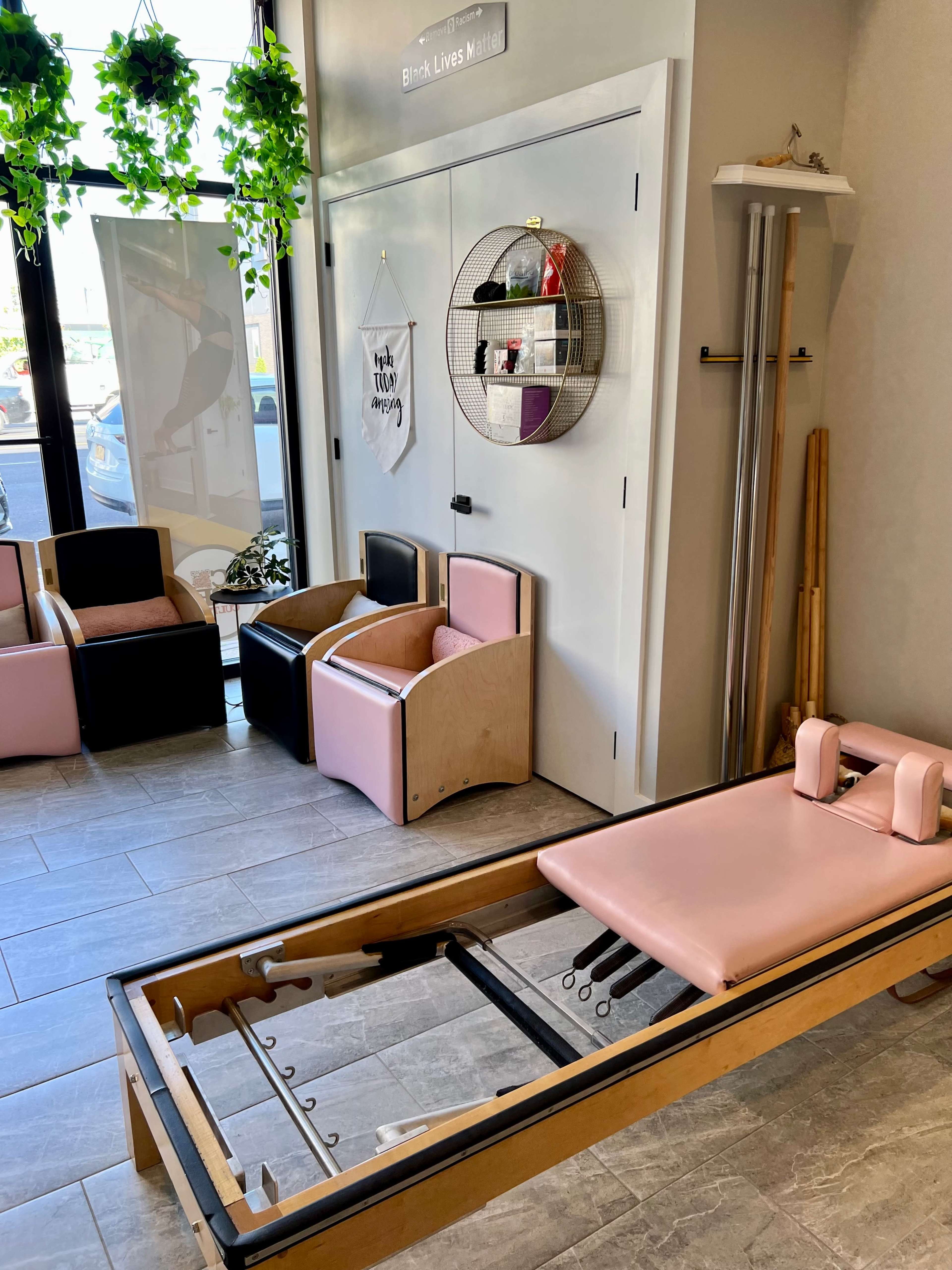 A wellness studio with a pink reformer machine, various seating options, and a wall decorated with greenery and shelves.