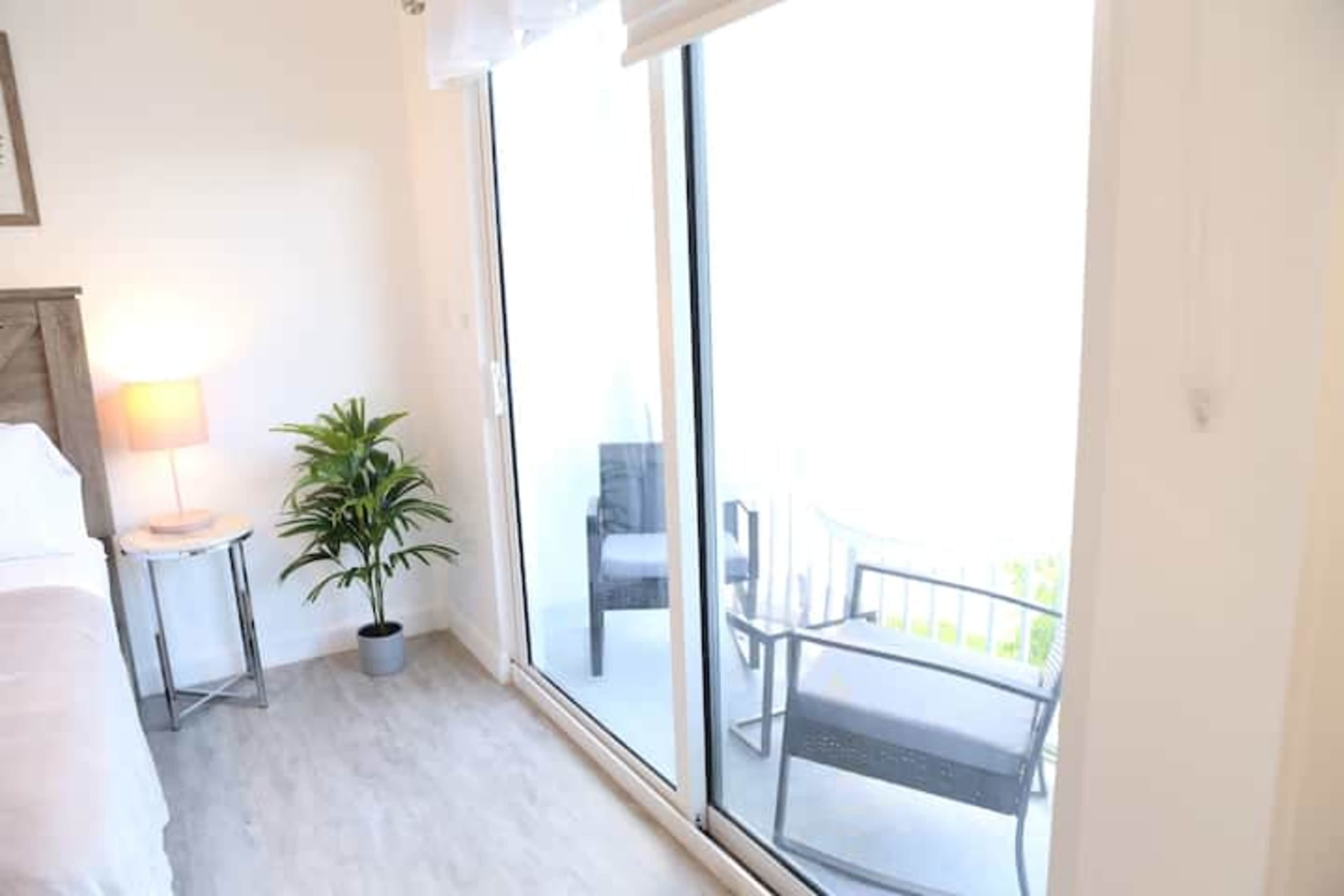 The image shows a well-lit bedroom featuring a bed, a bedside lamp, a potted plant, and a sliding glass door leading to a balcony with outdoor seating.