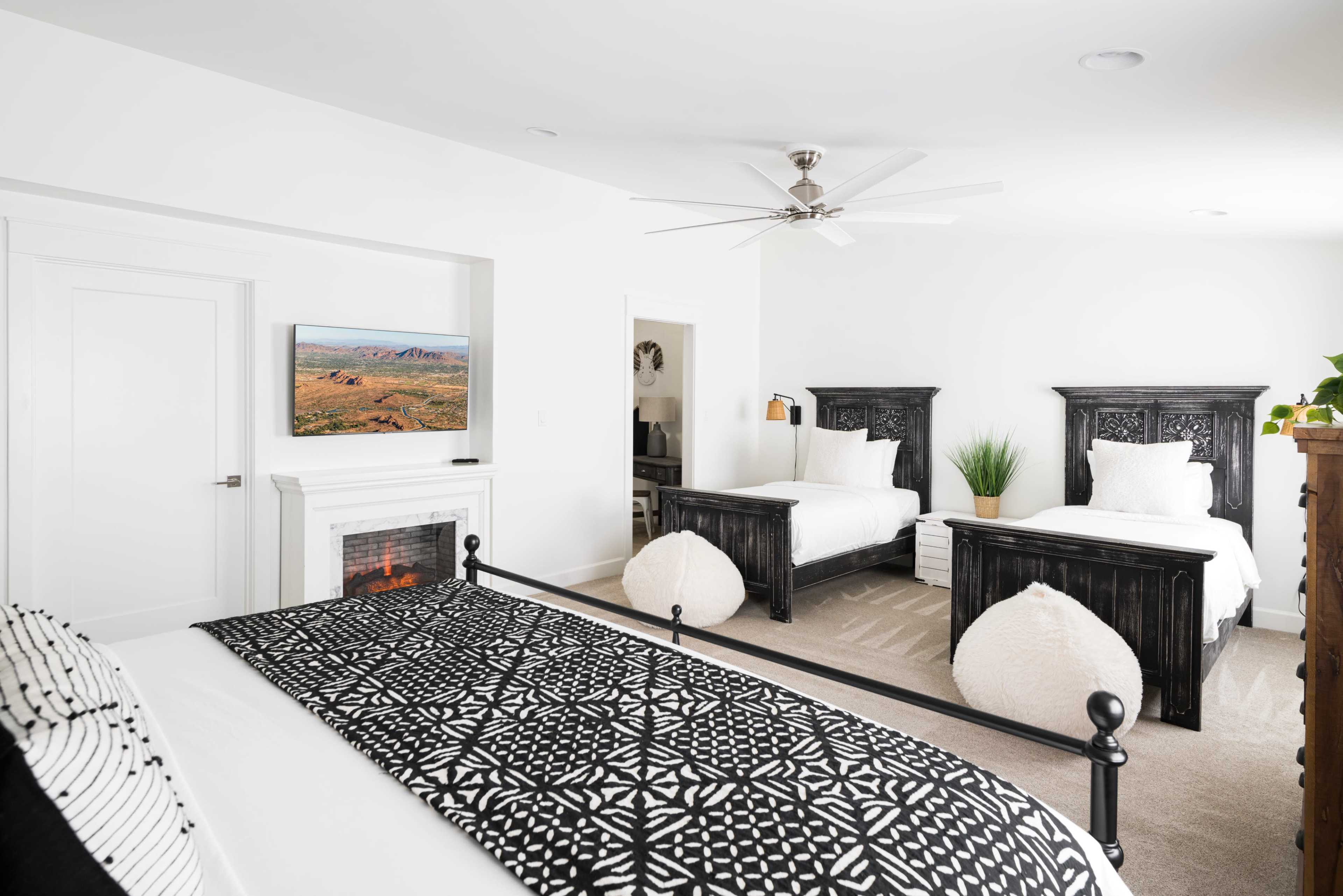 The image shows a well-lit bedroom featuring two twin beds with dark wooden headboards, a television mounted on the wall, and a cozy seating area.