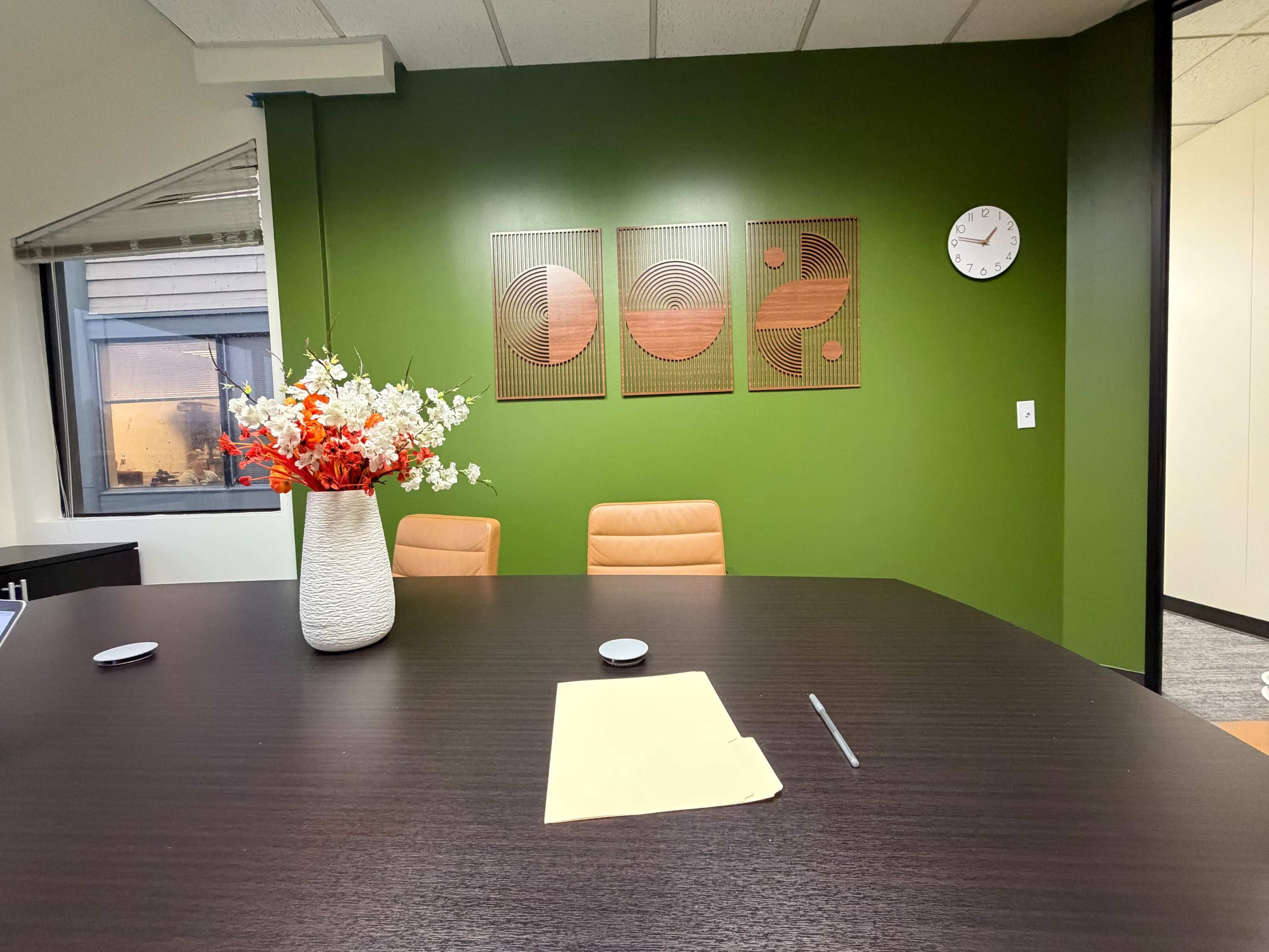The image shows a modern conference room with a dark wooden table, two orange chairs, a decorative vase with flowers, and three abstract wall art pieces on a green wall.