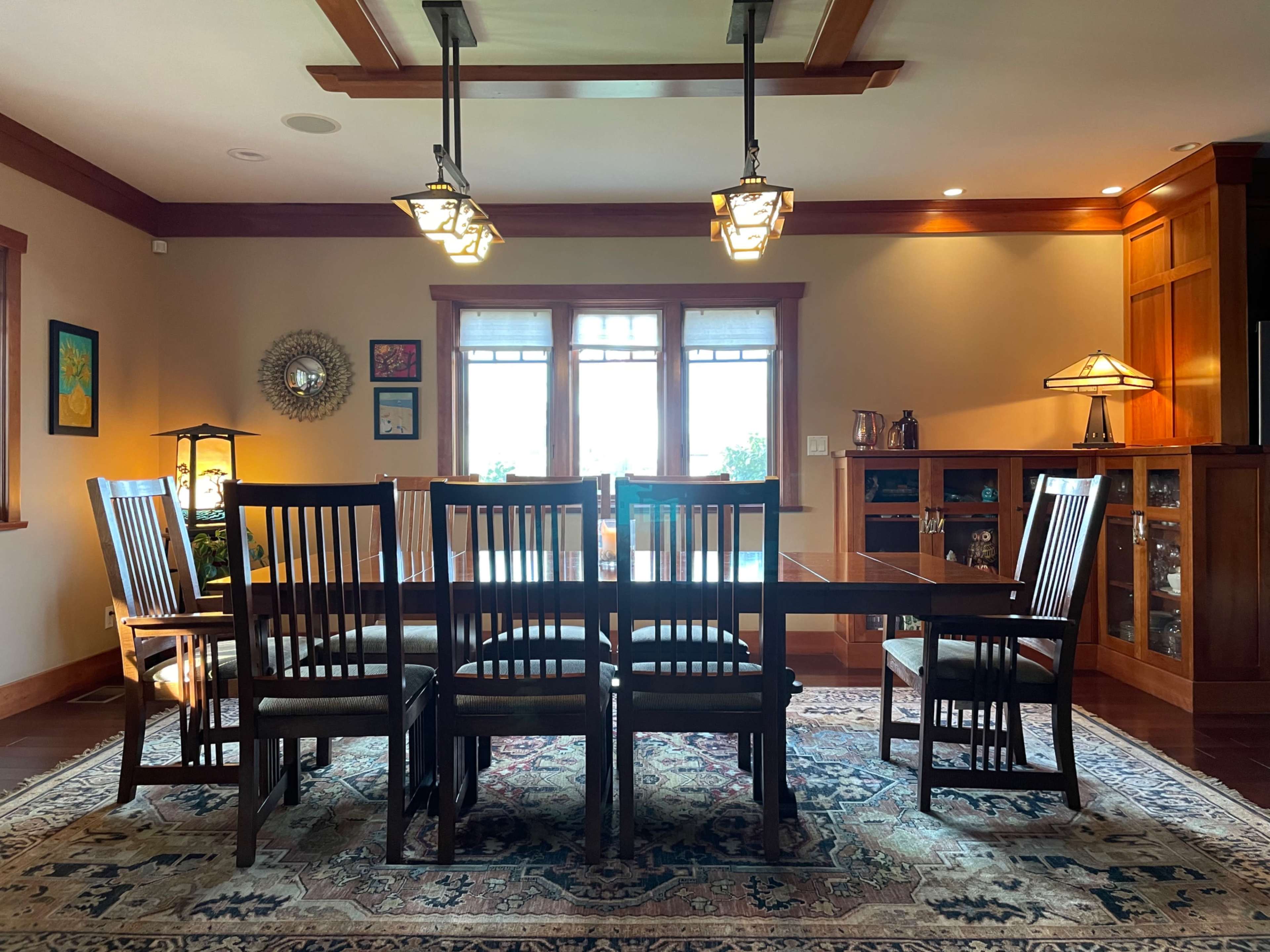 A spacious dining room features a long wooden table surrounded by eight chairs, with large windows allowing natural light to illuminate the space.