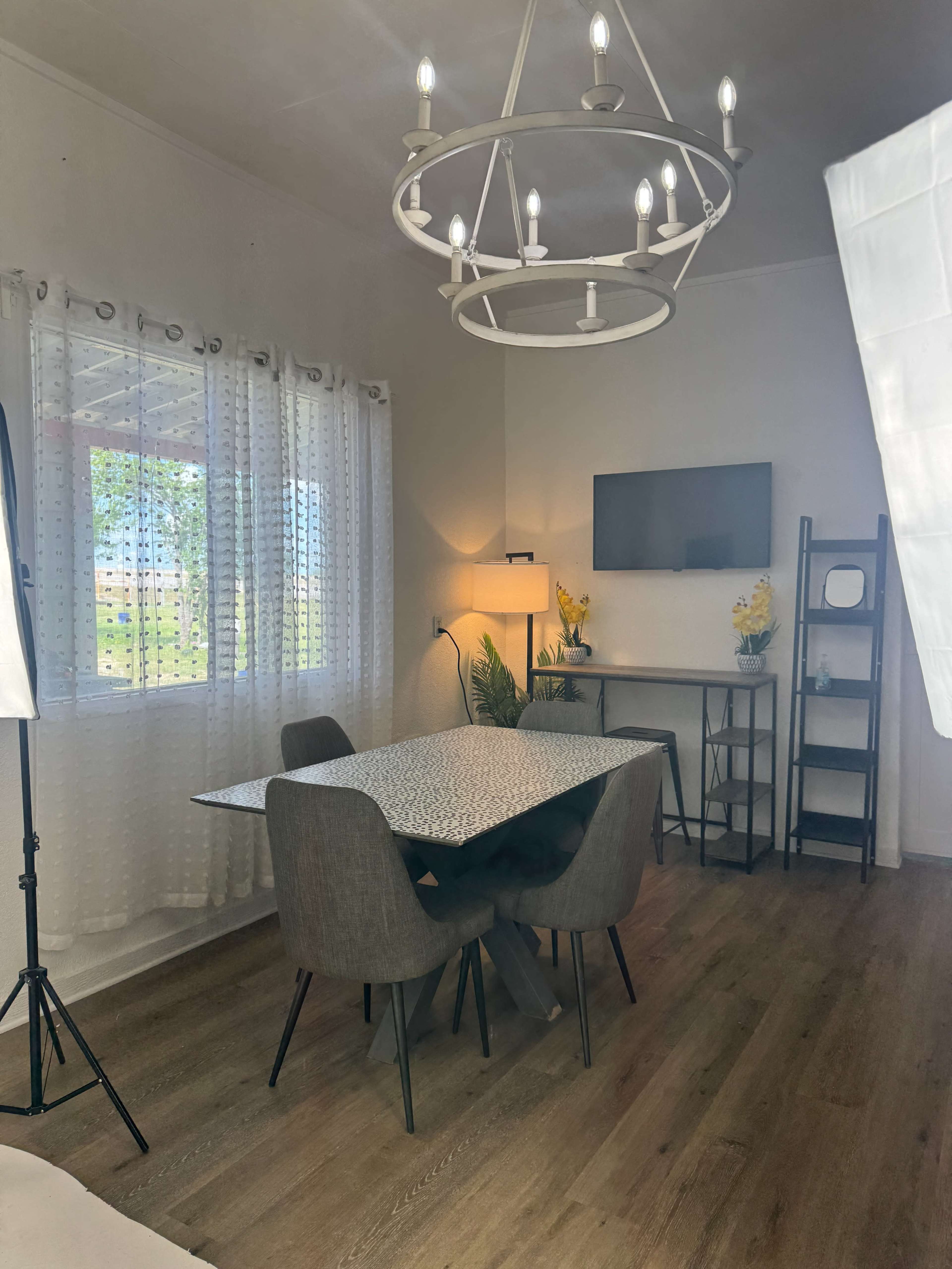 A modern dining area features a table with four chairs, a flat-screen TV on the wall, and a hanging chandelier, with natural light streaming through a window adorned with sheer curtains.