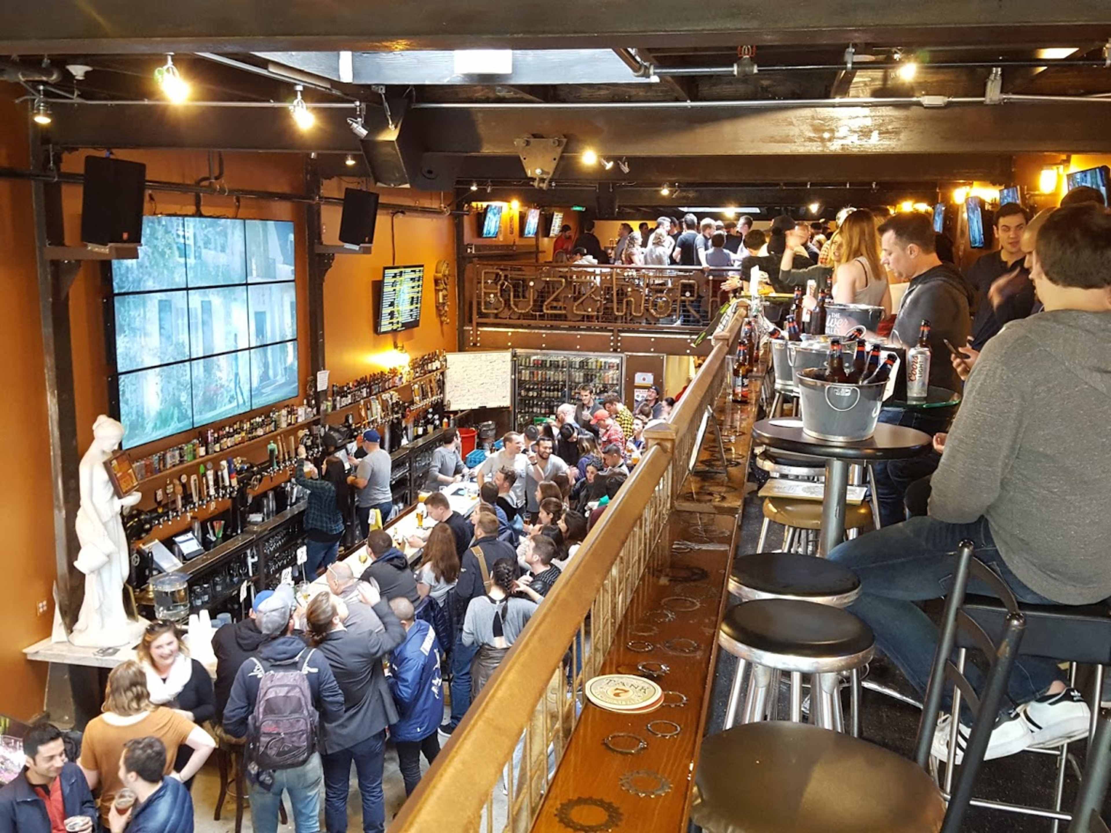 The image shows a busy bar filled with people, featuring a long counter with stools and shelves stocked with various beverages.