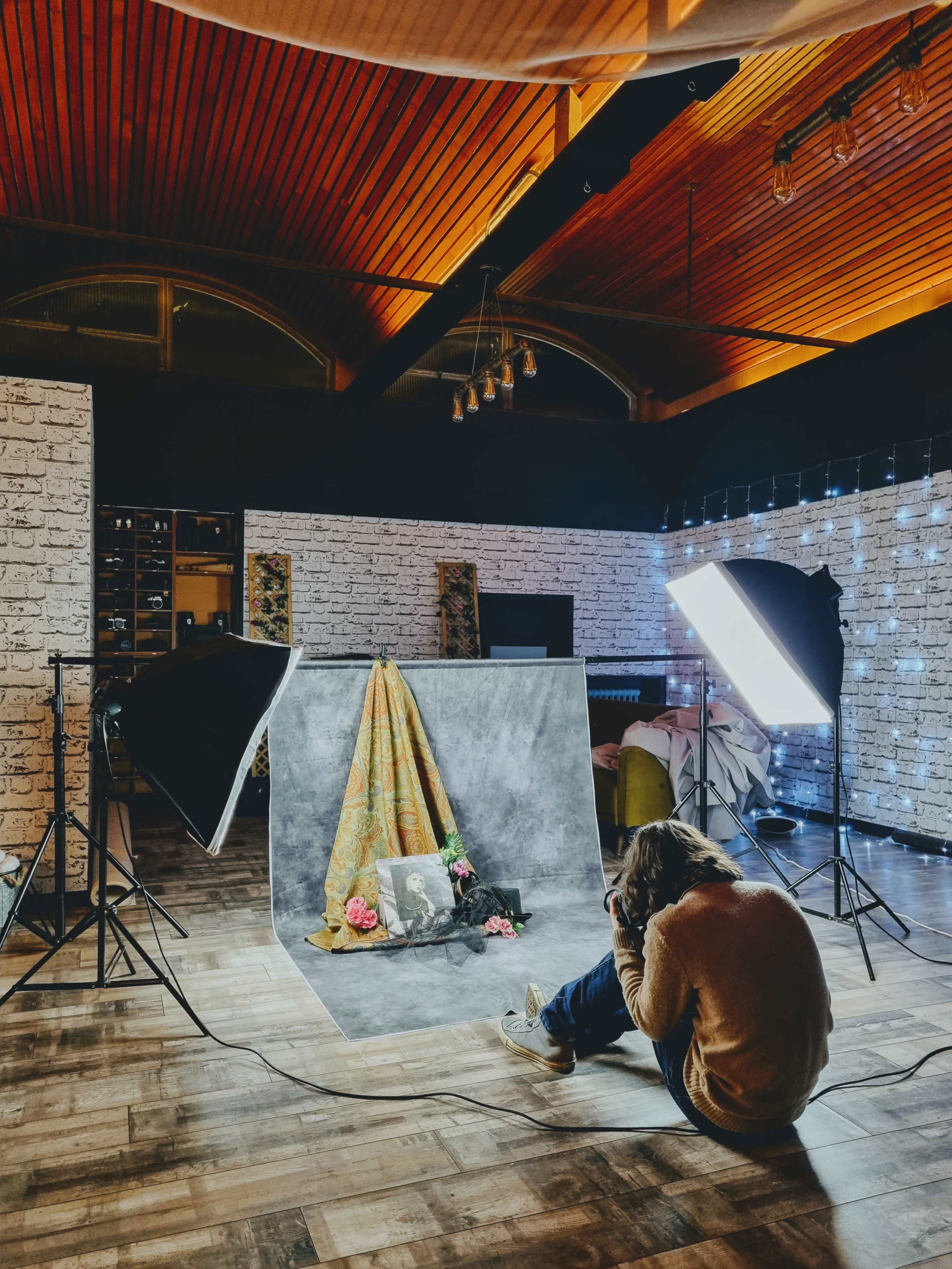 A photographer kneels in a studio, capturing an image of a decorated backdrop featuring fabric and flowers, with studio lights positioned to illuminate the scene.
