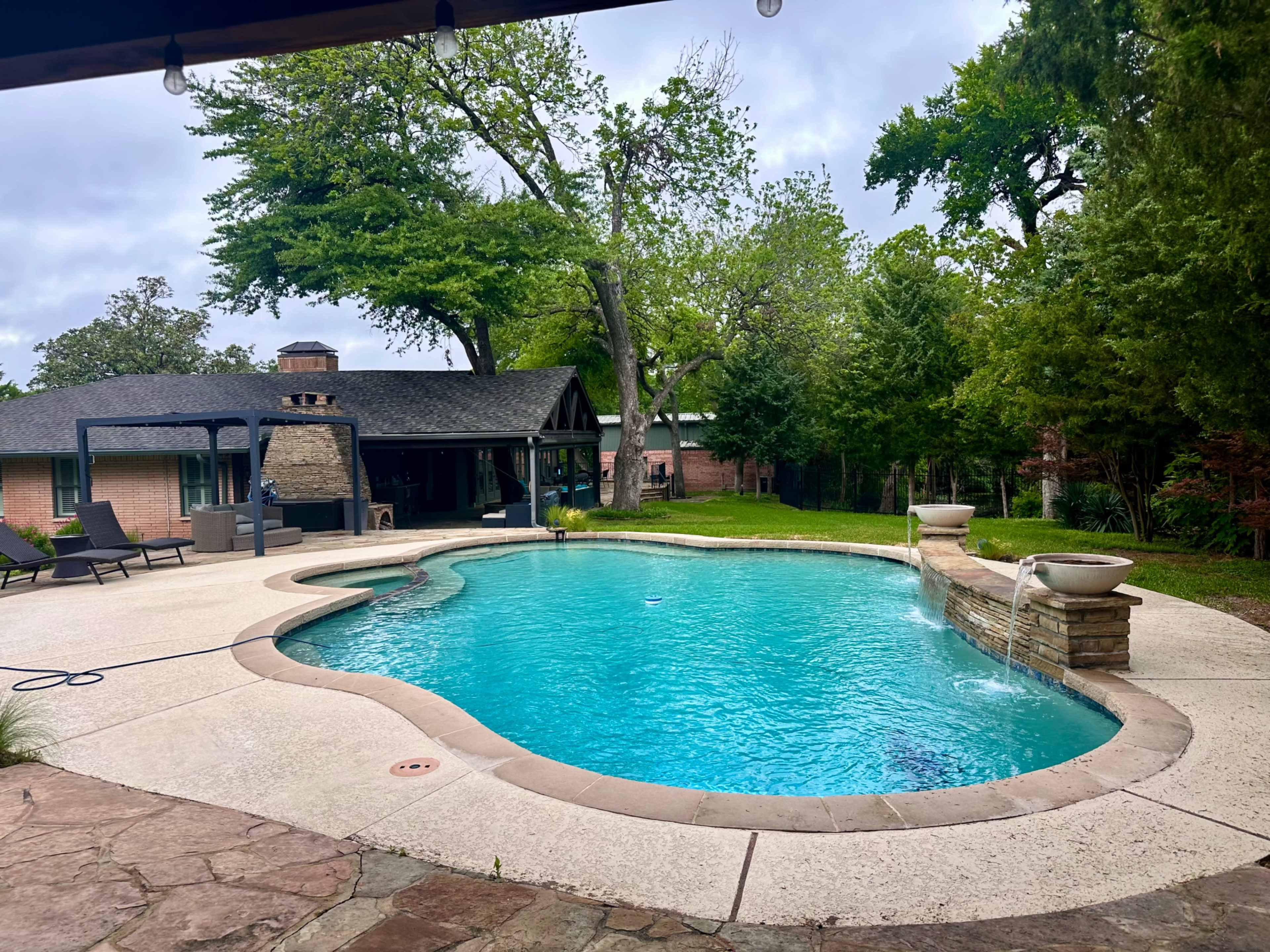 The image shows a backyard with a swimming pool, surrounded by greenery and a covered patio area.