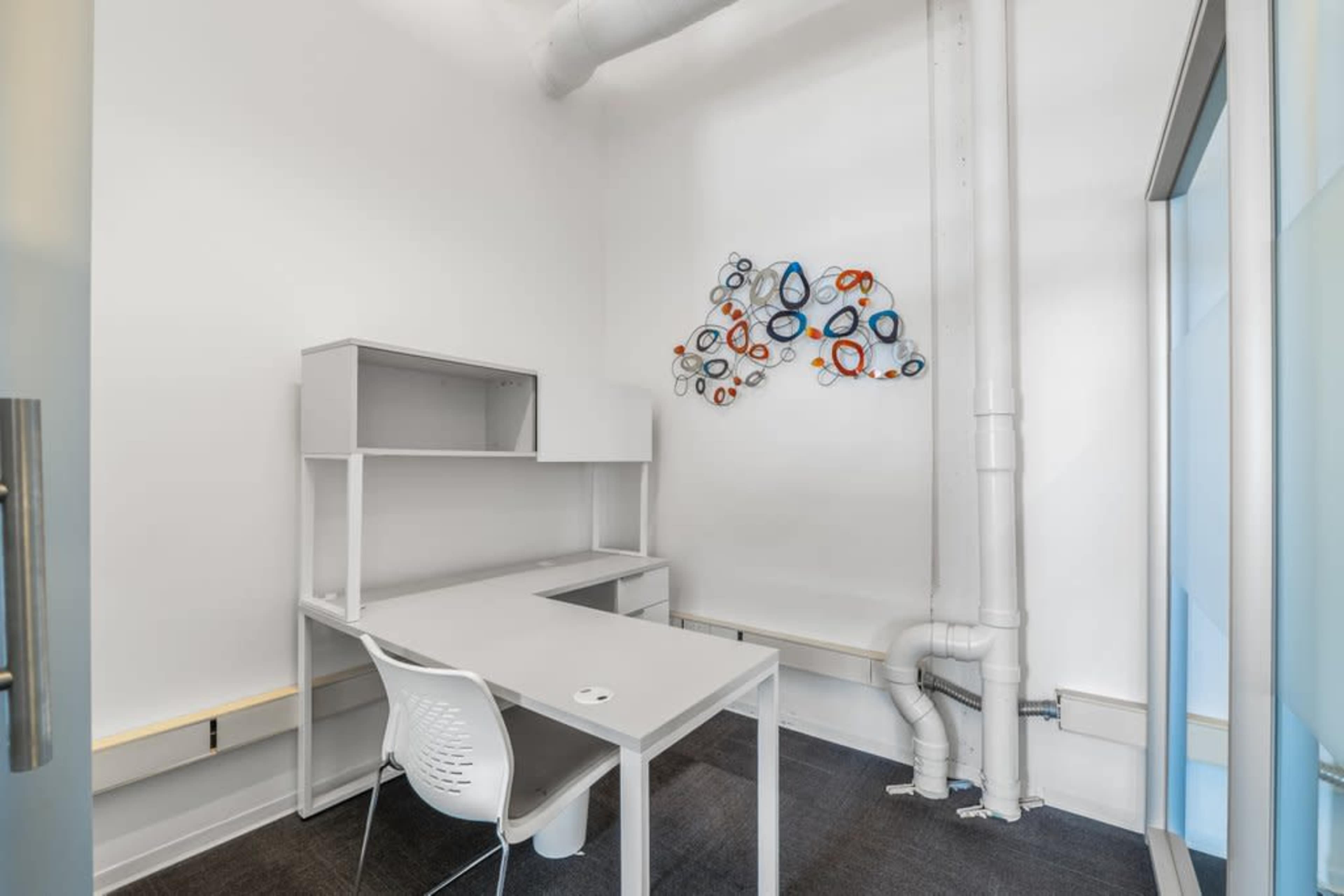 The image shows a minimalist office space featuring a gray desk and chair against a white wall, with abstract art made of colorful rings displayed above the desk.