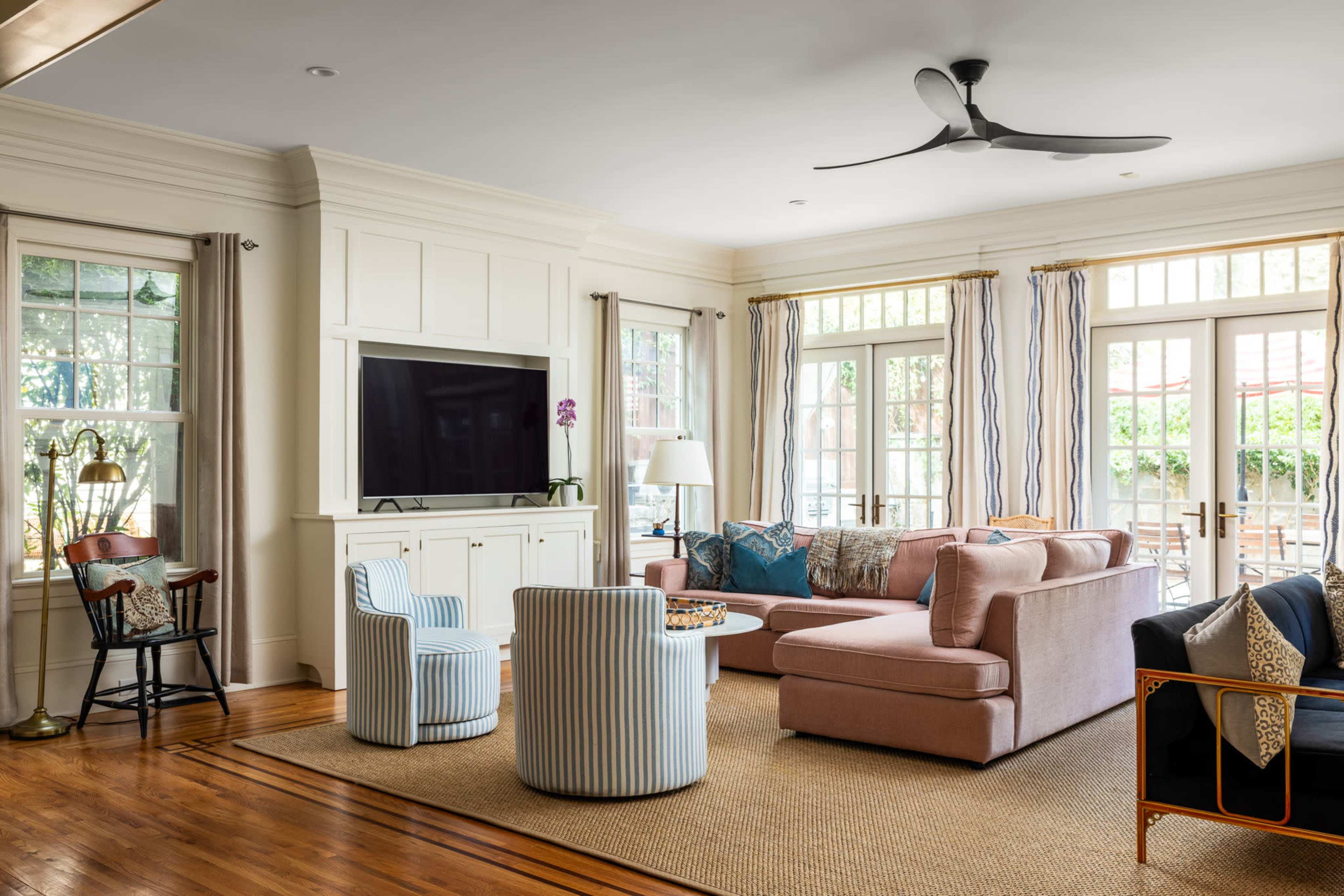 The living room features a pink sectional sofa, two striped chairs, a large television, and large windows that open to a view of the outdoor space.