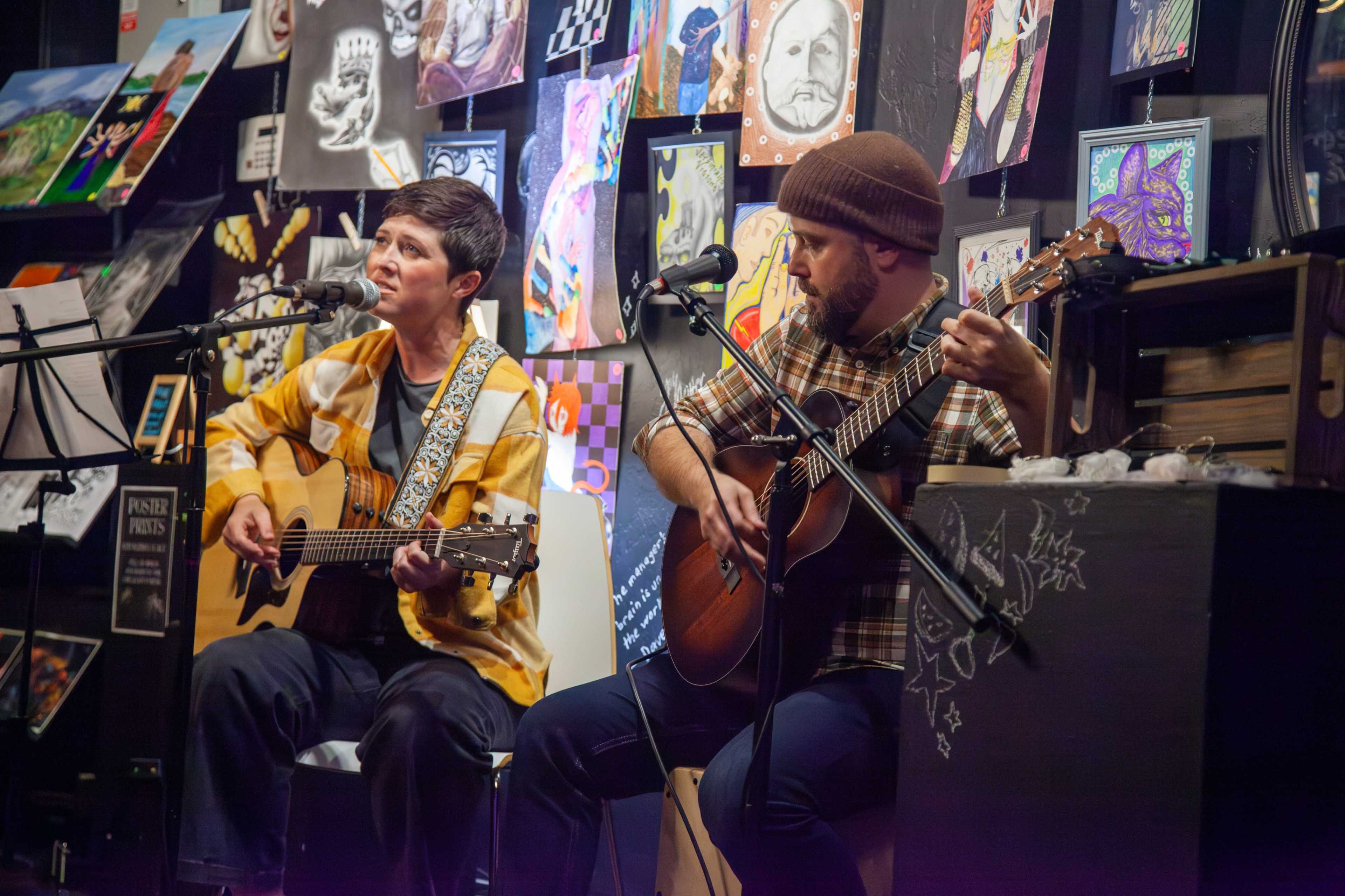 Two musicians perform live on stage, with one playing an acoustic guitar and the other using a guitar, surrounded by an array of colorful artwork on the walls.