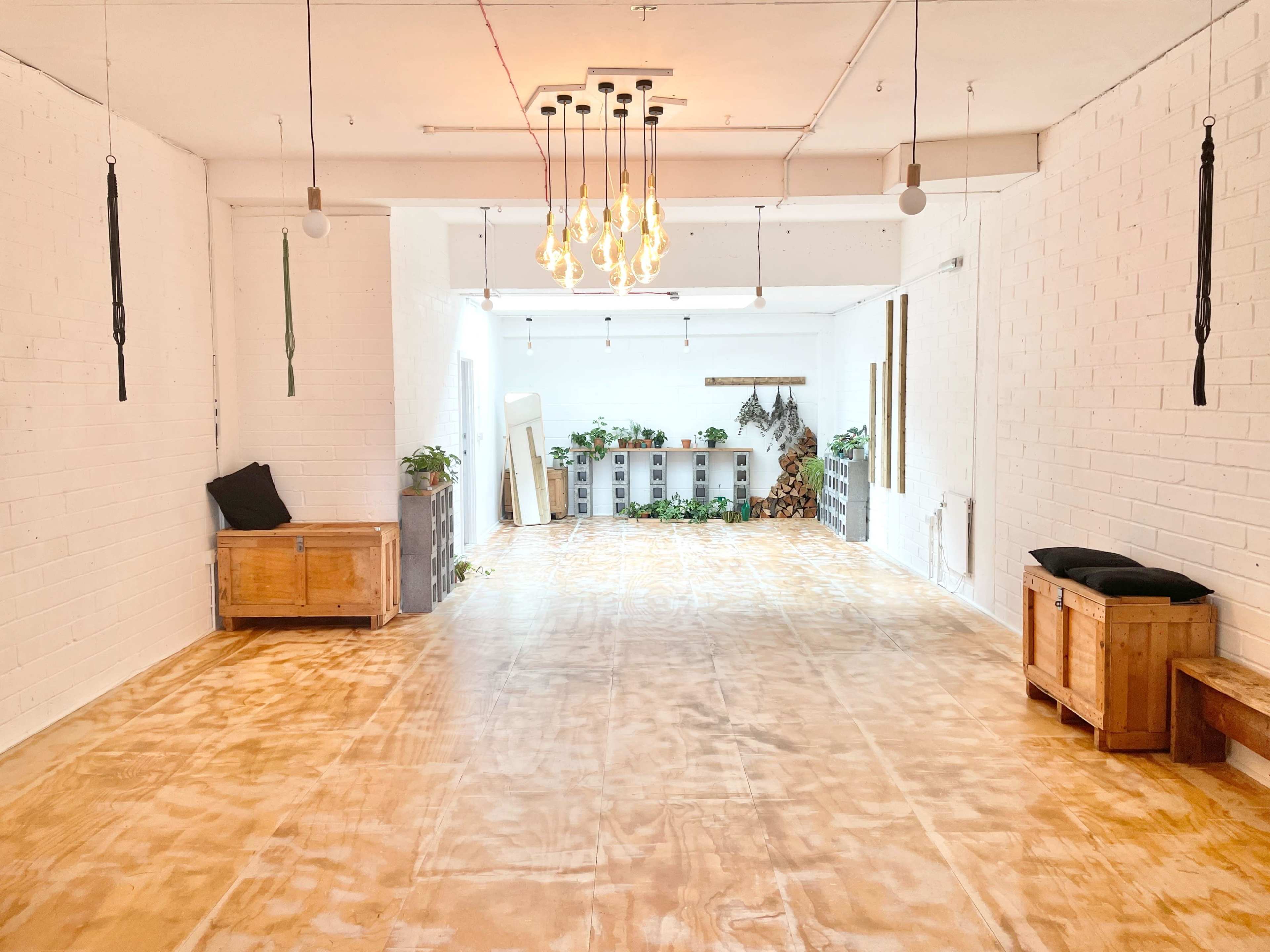 The image shows a spacious, bright room with wooden flooring, white brick walls, hanging light fixtures, and various plants, along with wooden furniture arranged along the sides.