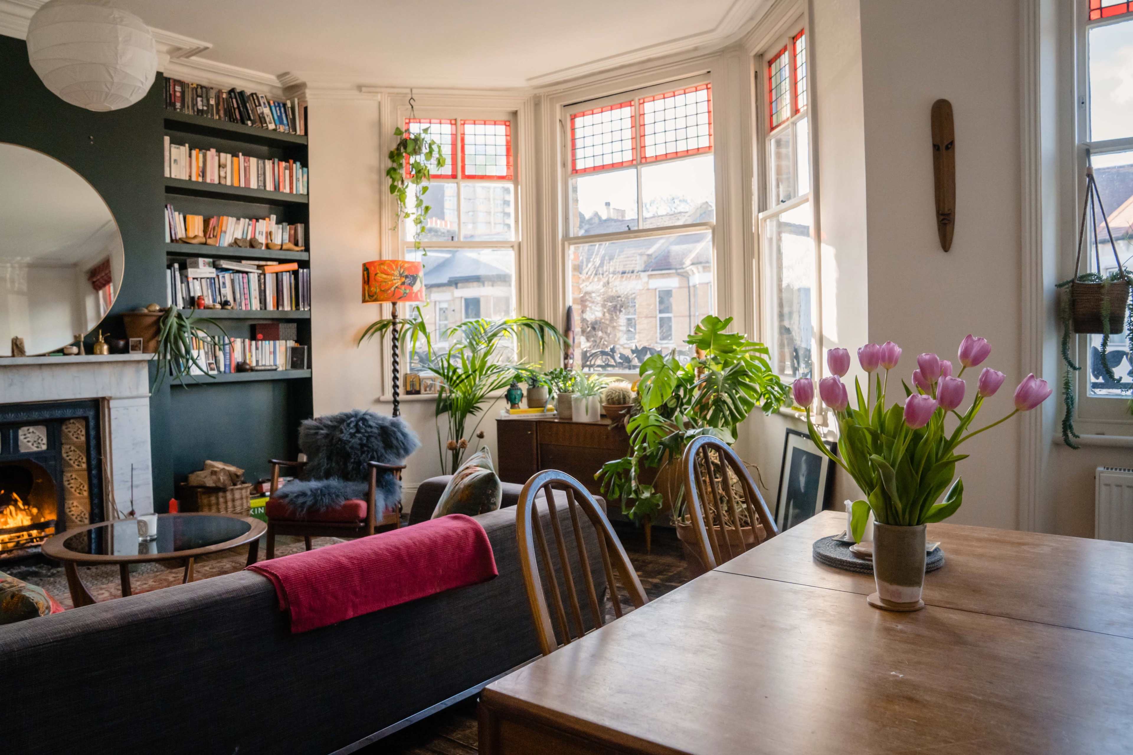 A cozy living room features a bookshelf, a dining table, and large windows with plants and flowers creating a welcoming atmosphere.