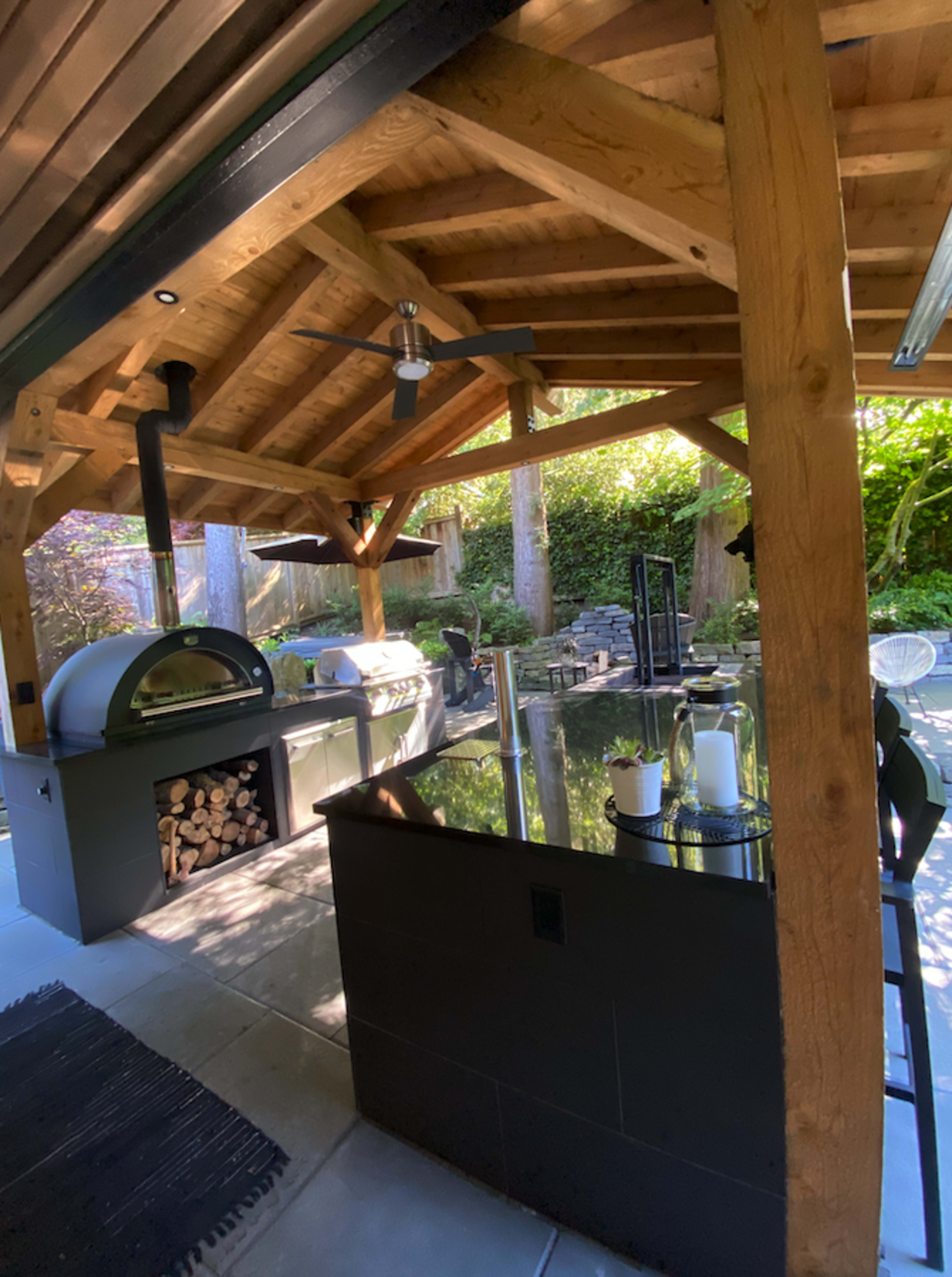 The image shows an outdoor kitchen with a wood-fired pizza oven, a bar area, and wooden beams supporting a slanted roof in a landscaped setting.