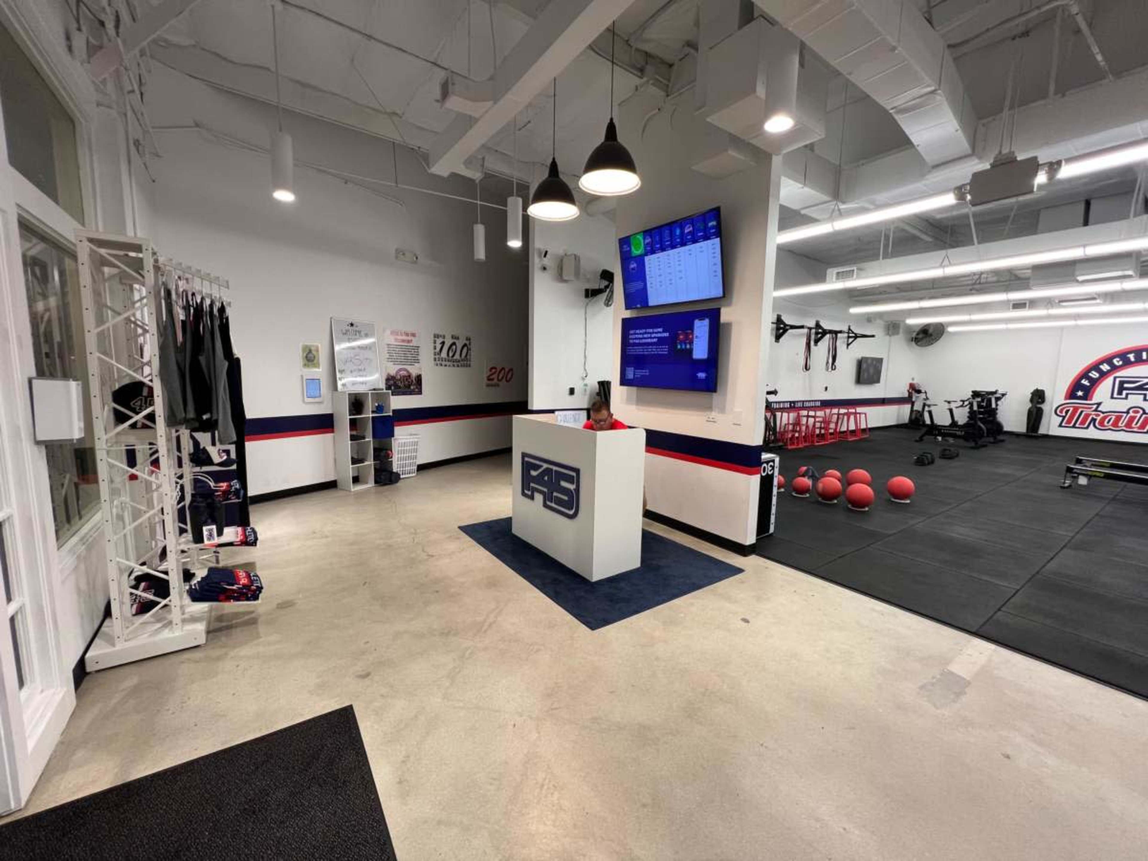 The image shows a modern fitness facility reception area with a white desk, a large screen displaying information, and workout equipment visible on the right side.
