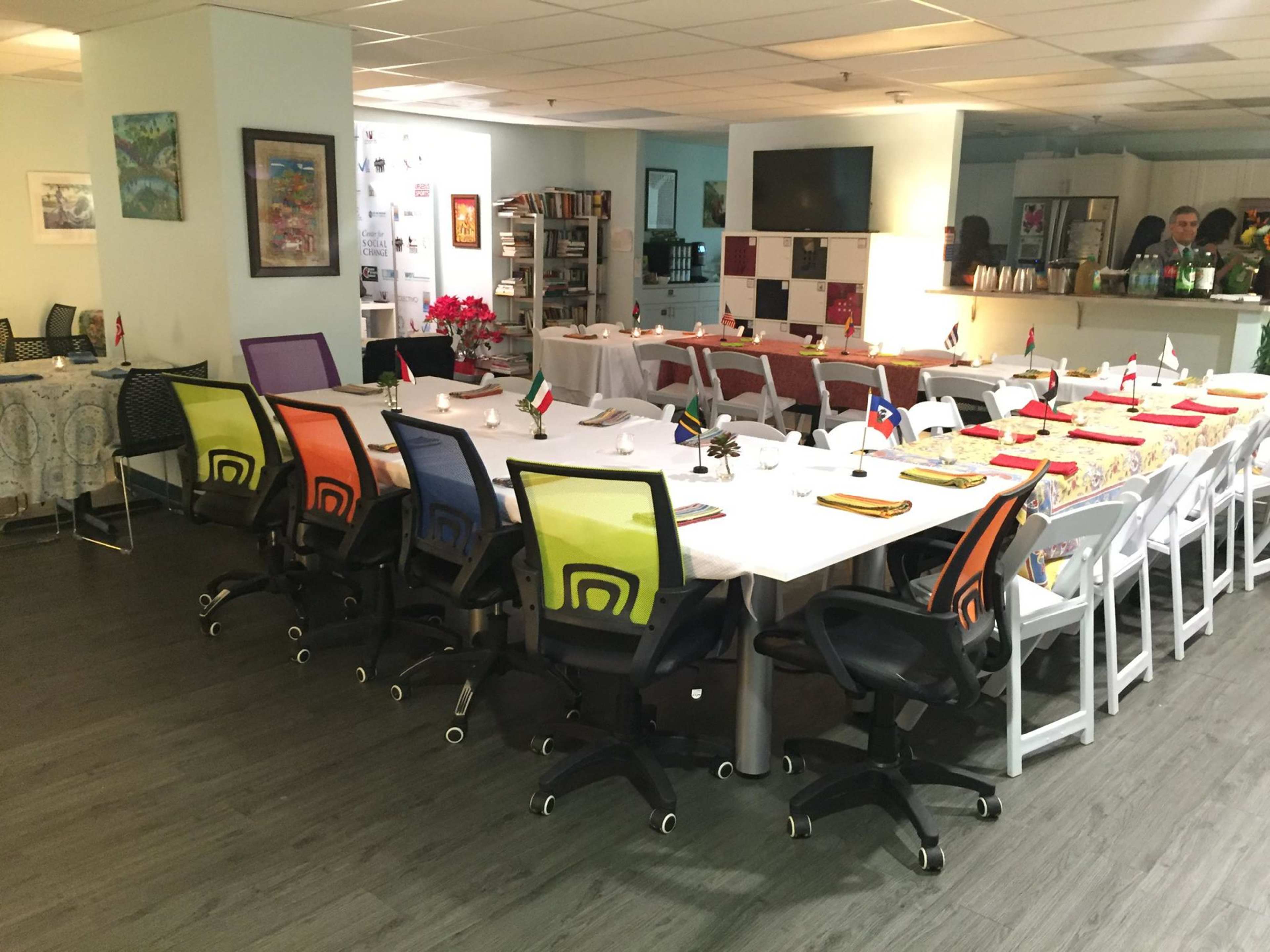 A large dining table with colorful chairs is set up in a well-lit room, surrounded by various decorations and flags from different countries.