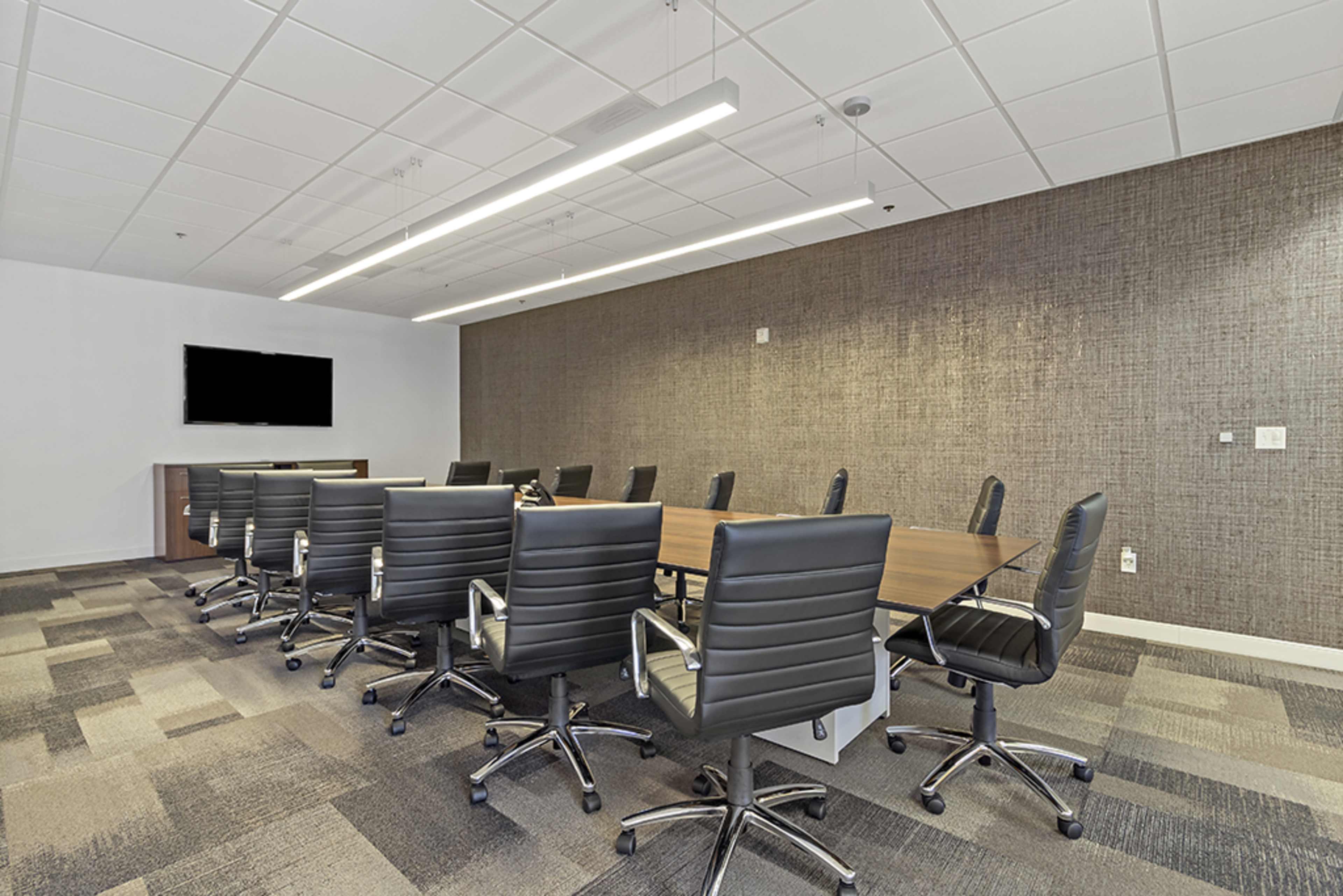 A large conference room features a long wooden table surrounded by black ergonomic chairs, with a wall-mounted screen at one end.