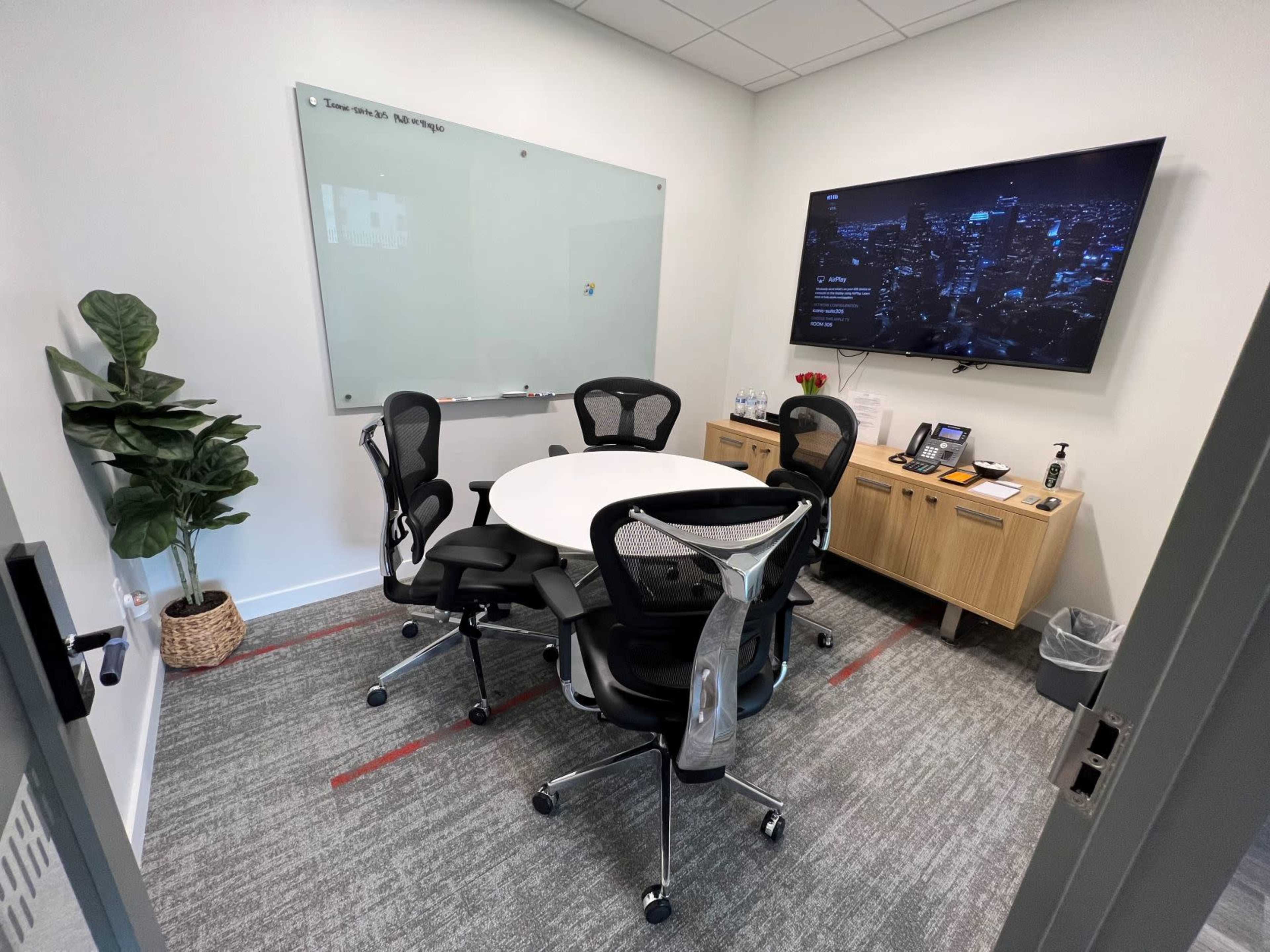 A small meeting room features a round table surrounded by six office chairs, a wall-mounted TV, a whiteboard, and a potted plant.