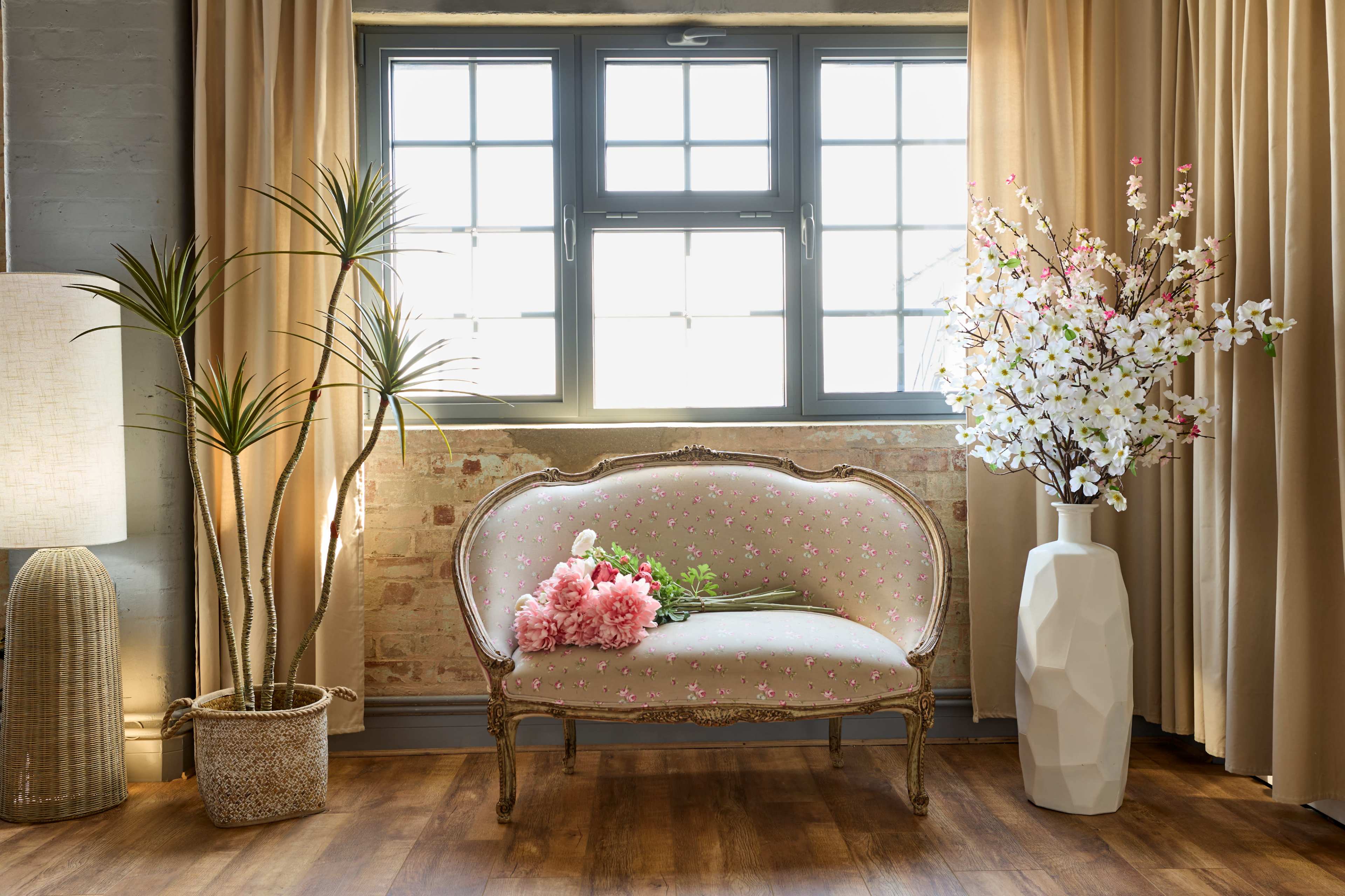 A floral-patterned settee is positioned near a window, accompanied by a tall plant and a large vase filled with artificial flowers.
