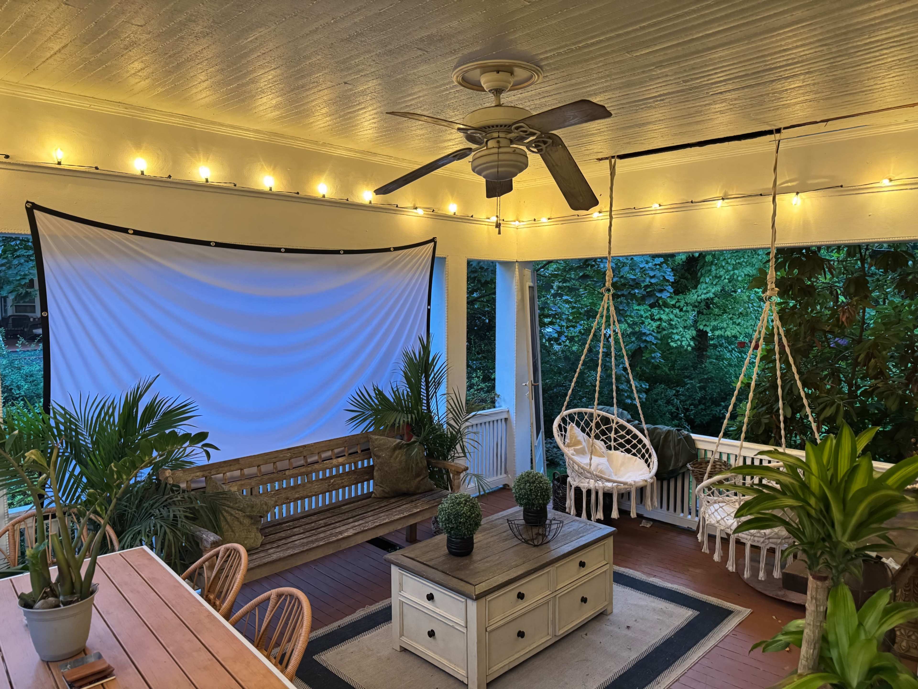The image shows a cozy outdoor patio area with a hanging ceiling fan, string lights, and two hanging chairs, surrounded by greenery.