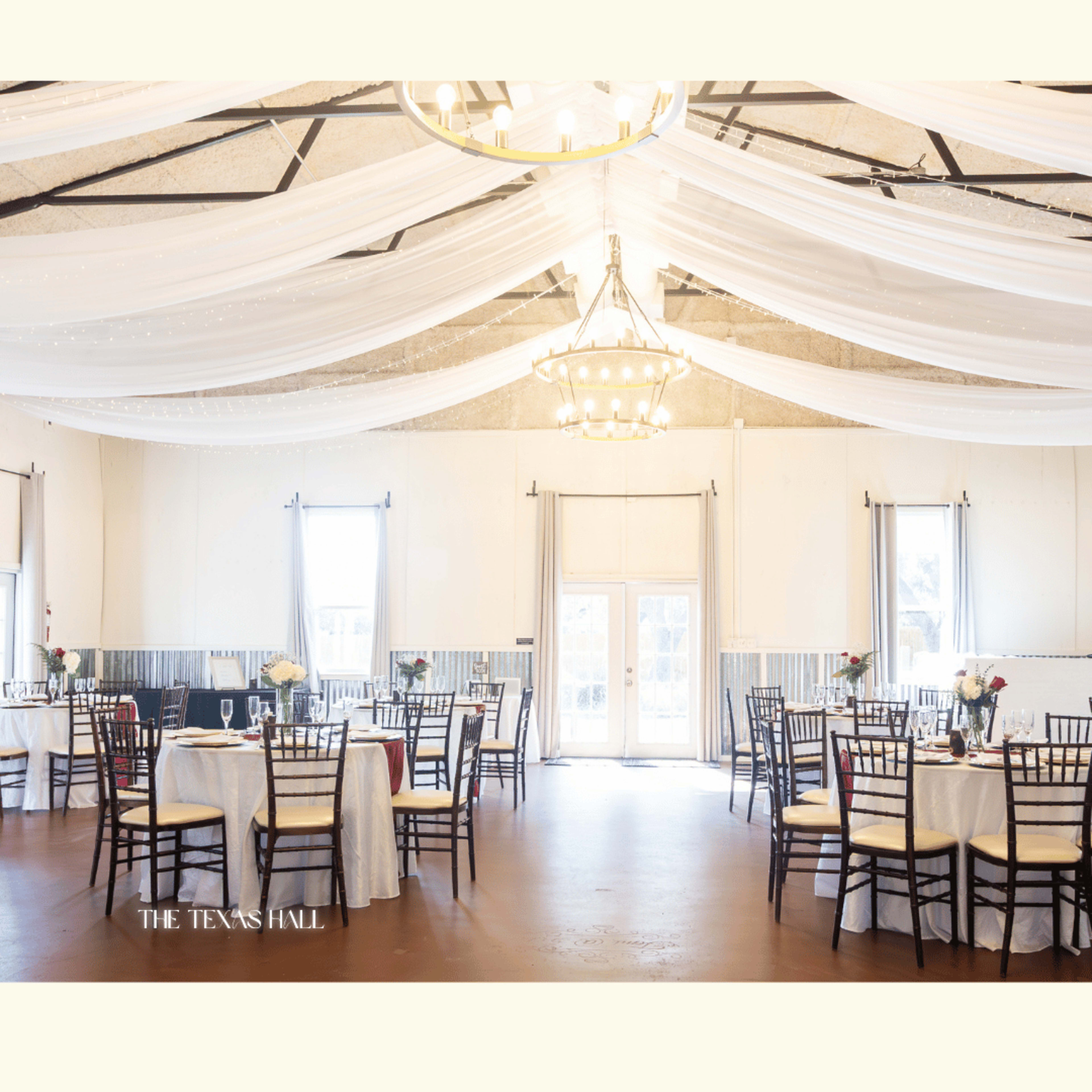 The image shows a spacious event hall with tables set for dining, draped fabric hanging from the ceiling, and a chandelier illuminated above.
