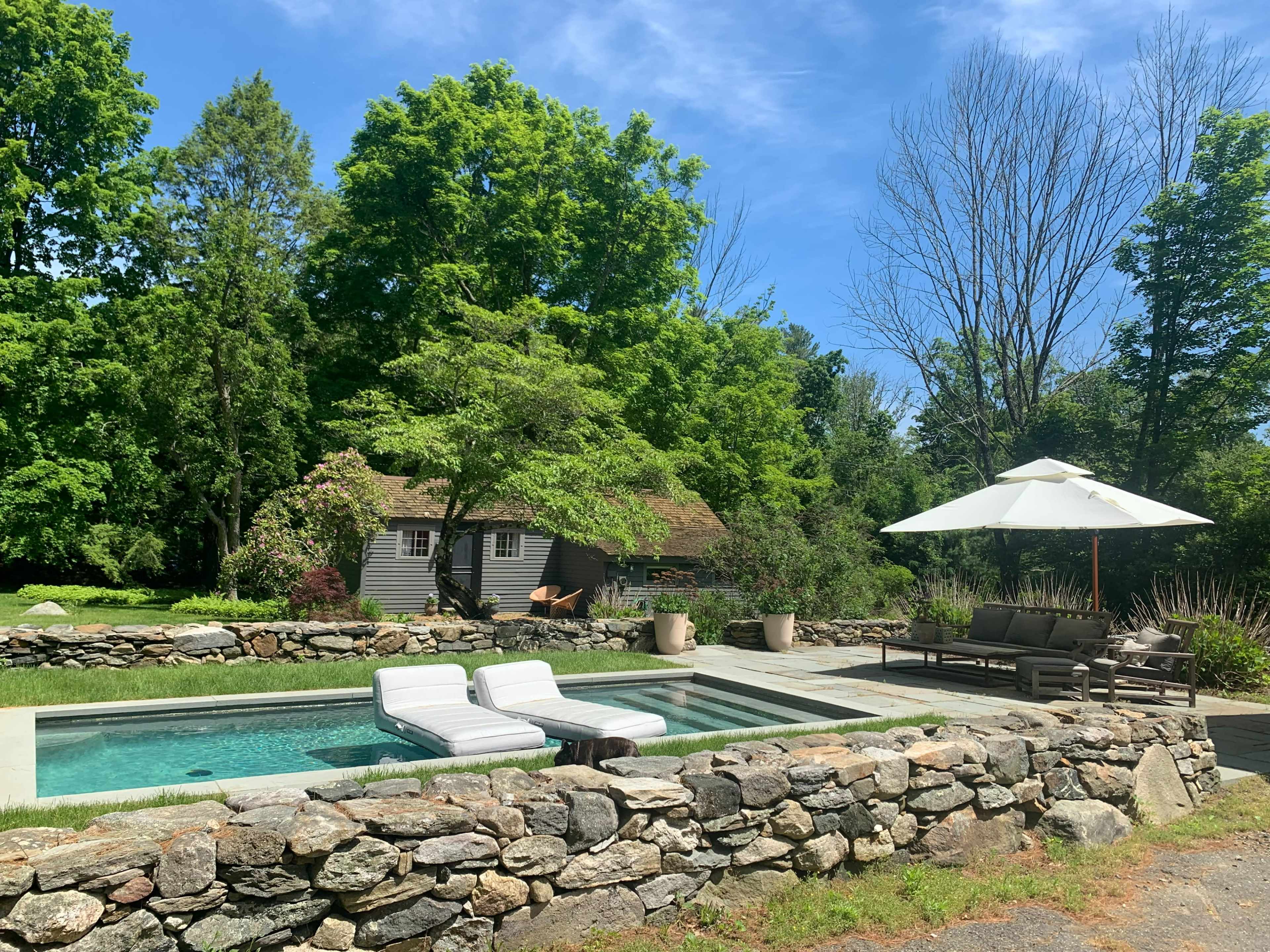 A rectangular pool with lounge chairs is surrounded by a stone wall and greenery, with a shaded seating area and a rustic building in the background.