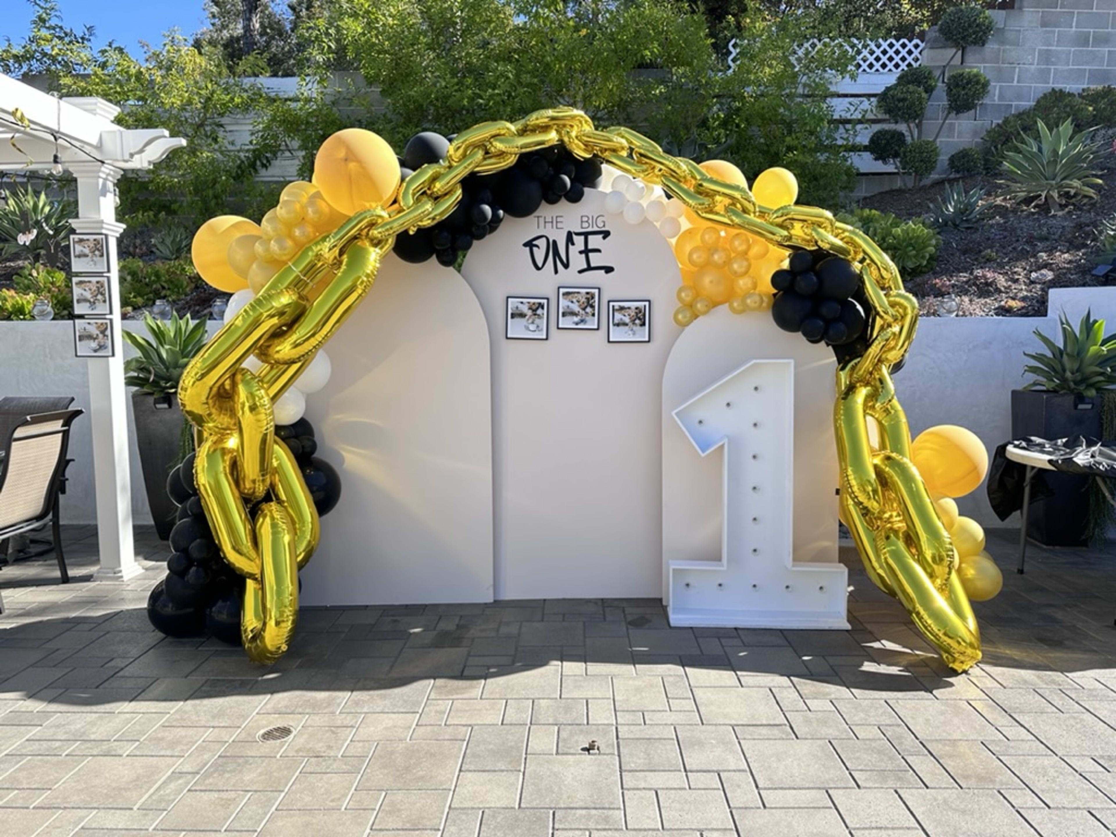 A festive birthday decoration featuring a large gold "1" and a balloon arch made of black, gold, and white balloons, set against a white backdrop with framed photos.