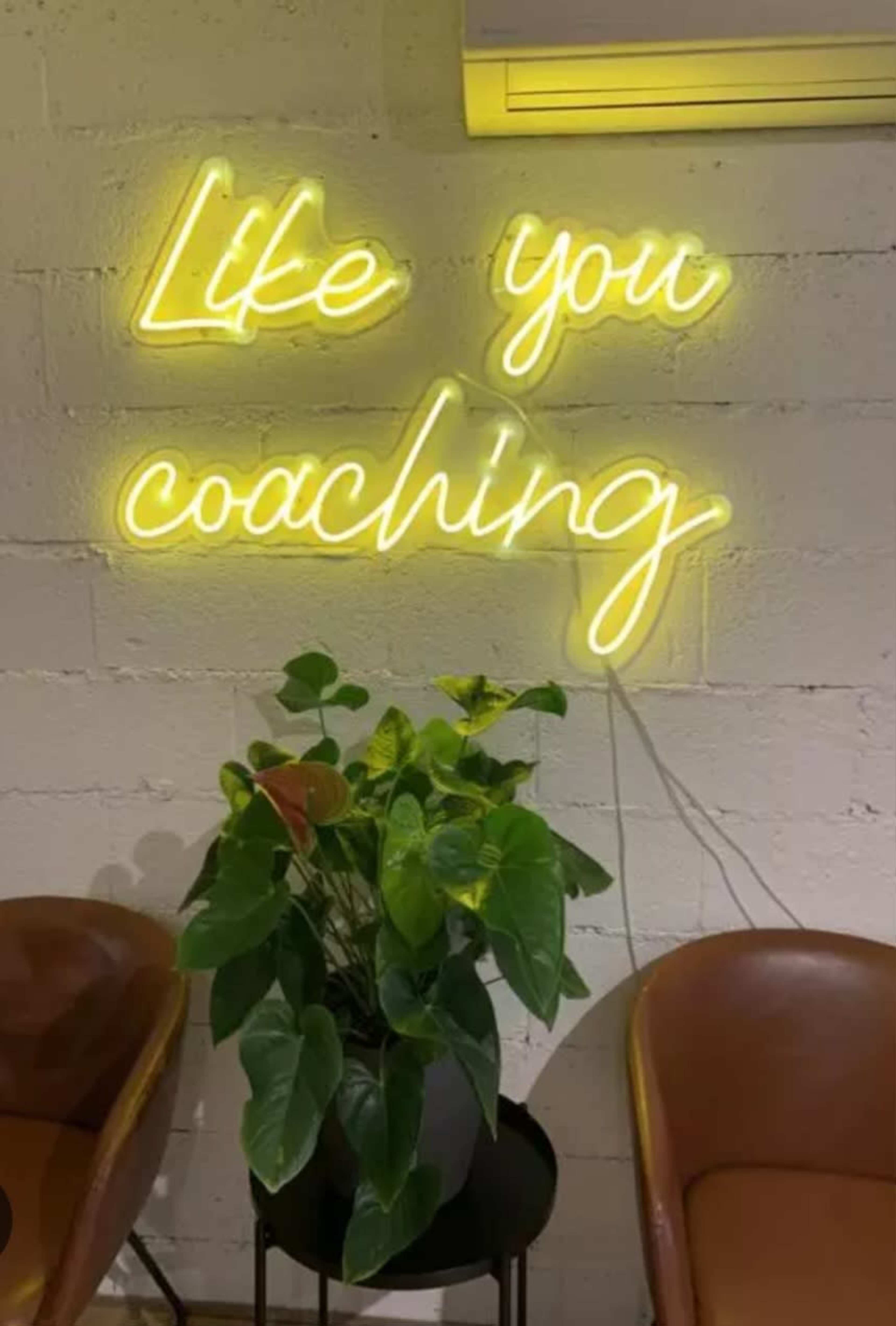 A neon sign displaying the words "Like you coaching" is positioned above a green plant on a small table, with two brown chairs in the foreground.
