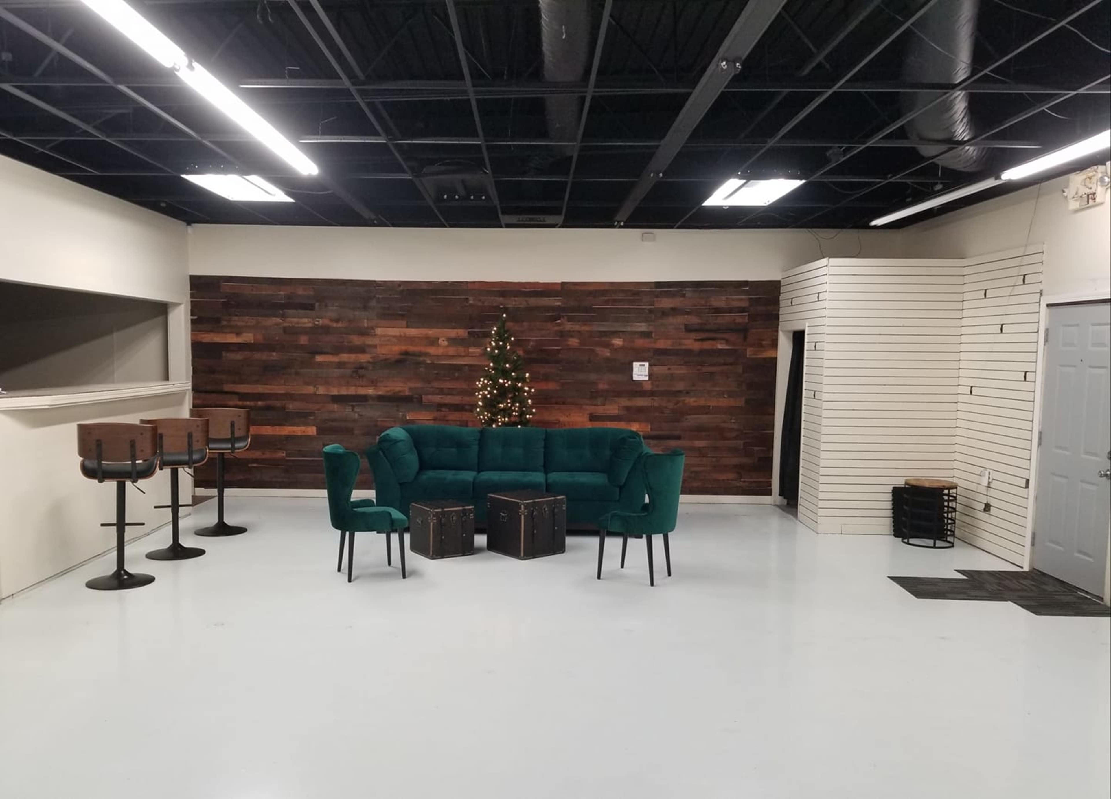 A green sectional sofa is arranged with two chairs and wooden crates in a spacious room featuring a wood-paneled wall and a small Christmas tree.