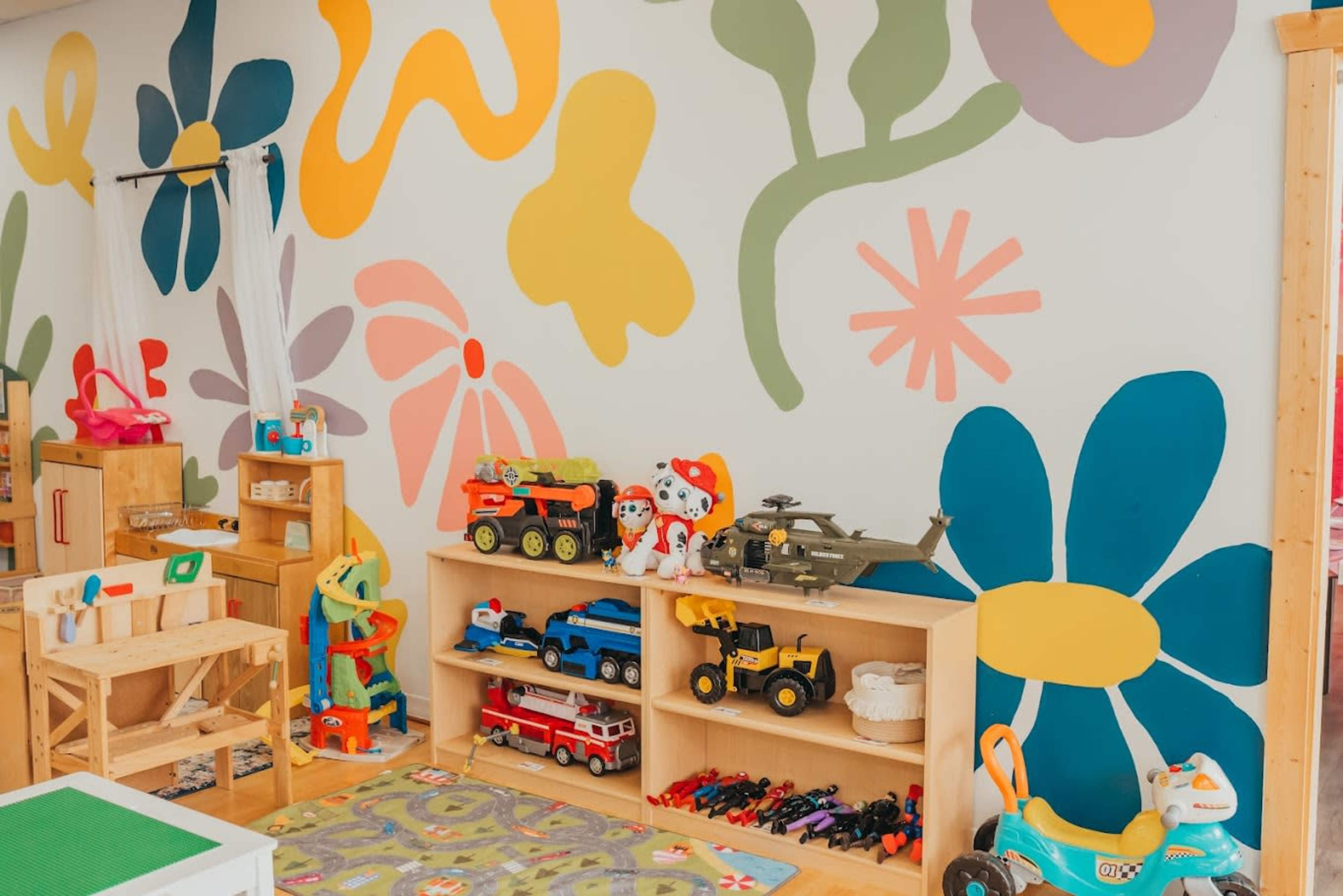 The image shows a brightly decorated room with a colorful mural on one wall and a shelf filled with various toys, including vehicles and action figures.