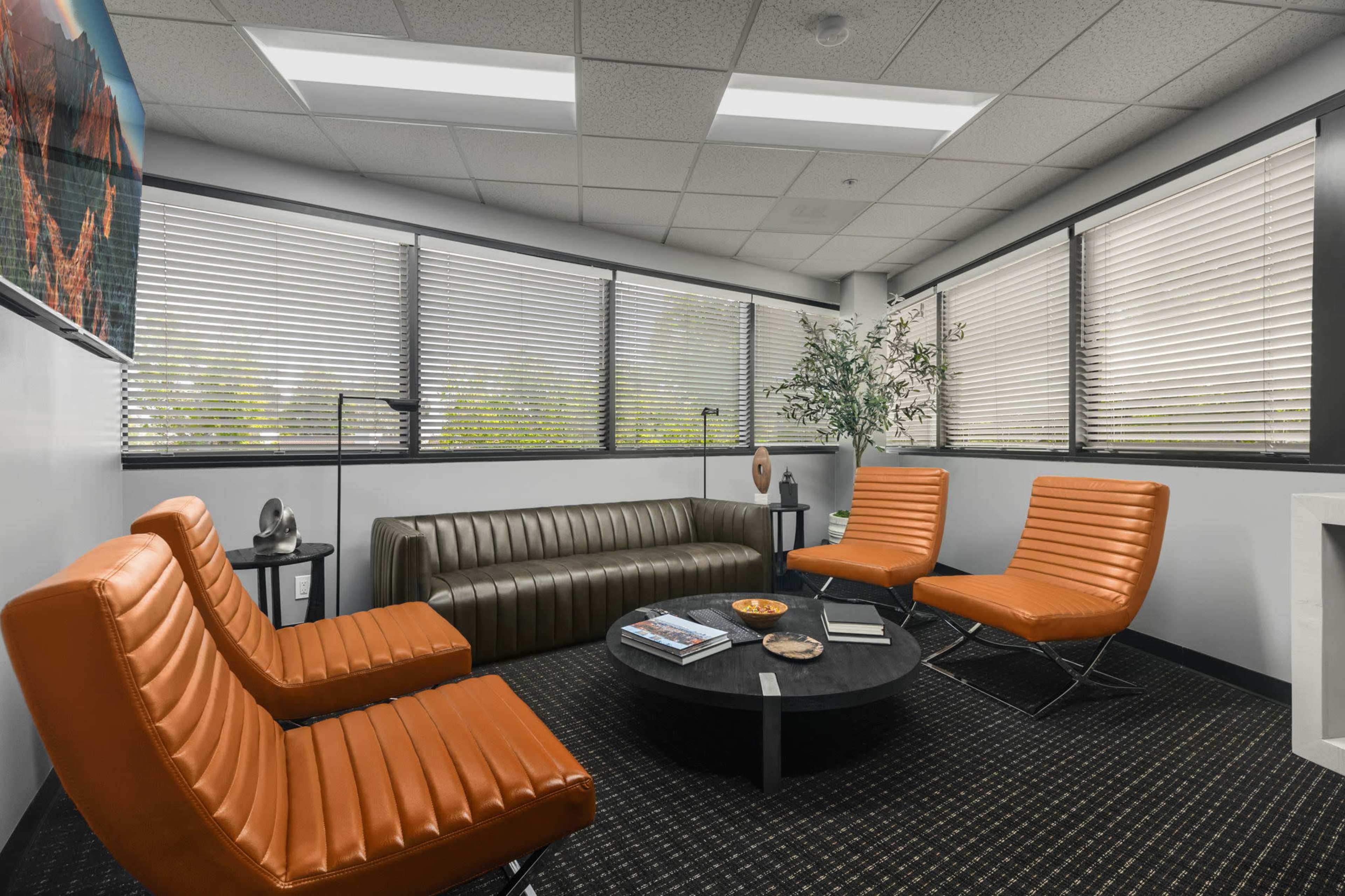 The image shows a modern office waiting area with a dark green leather sofa, two orange chairs, a round black coffee table, and large windows with blinds.