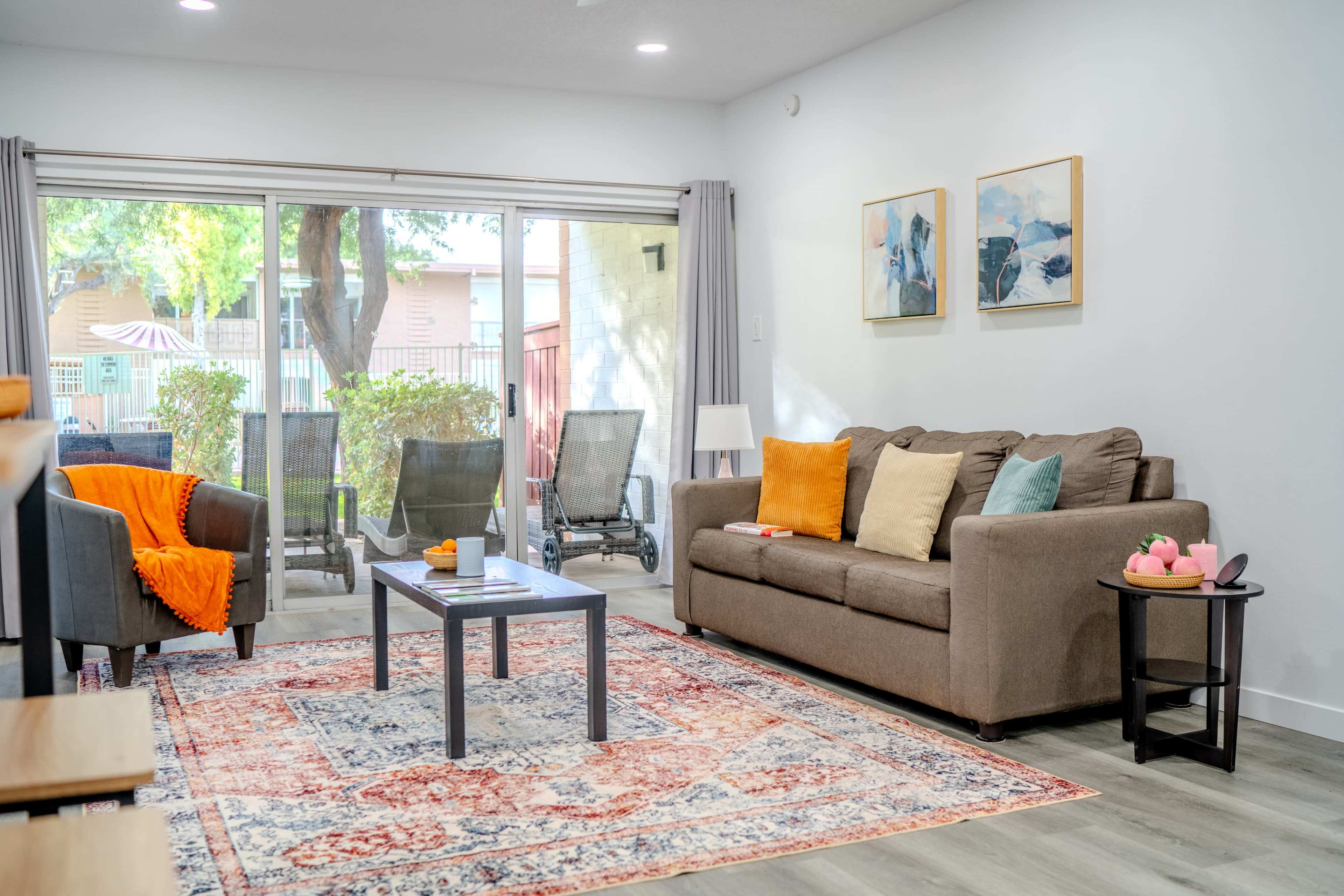 The image shows a modern living room with a brown sofa, two armchairs, a coffee table, and a large sliding glass door that opens to an outdoor patio area.