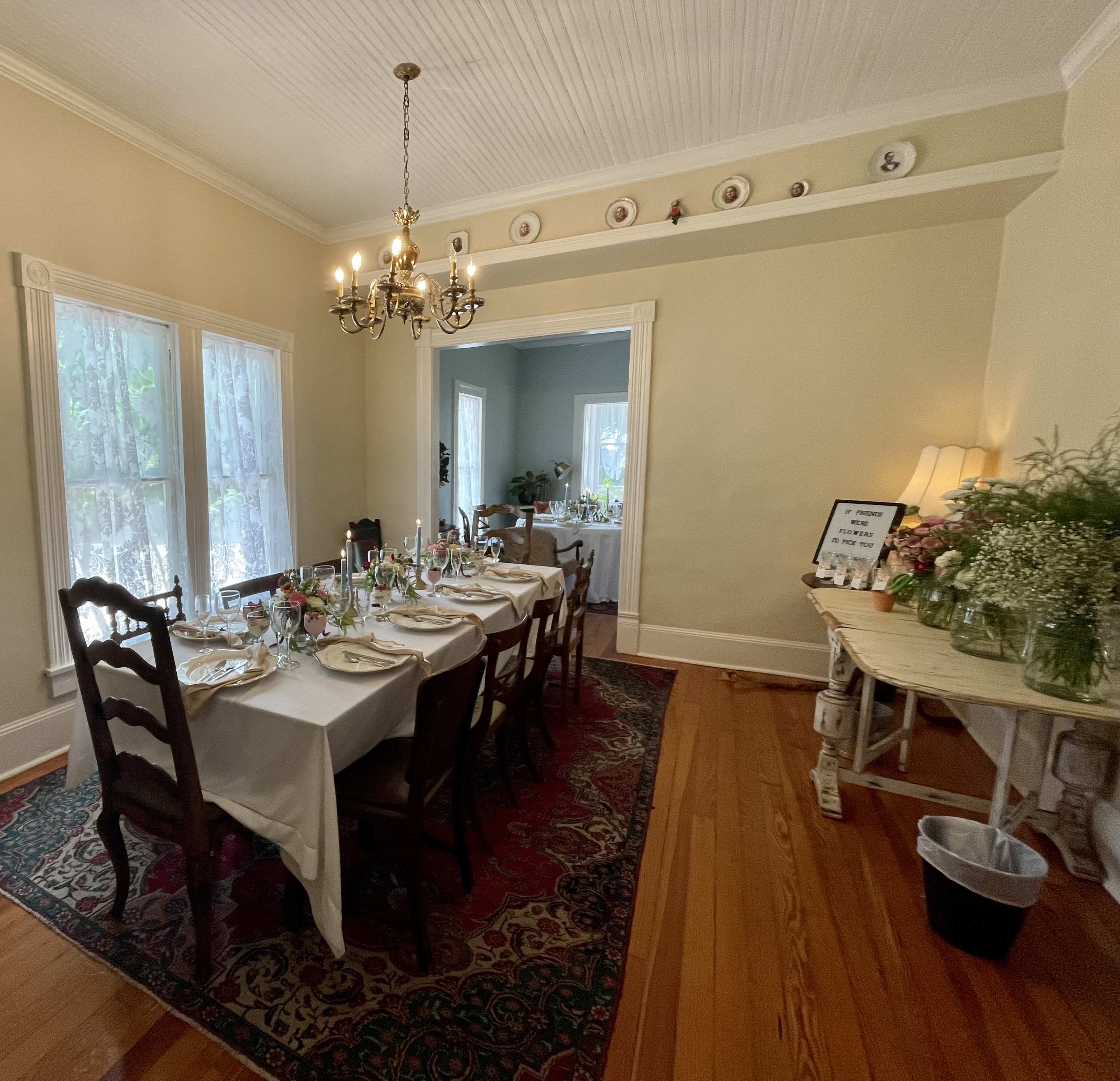 A dining room with a long table set for a meal, surrounded by wooden chairs, and featuring a chandelier overhead and decorative items on the walls.